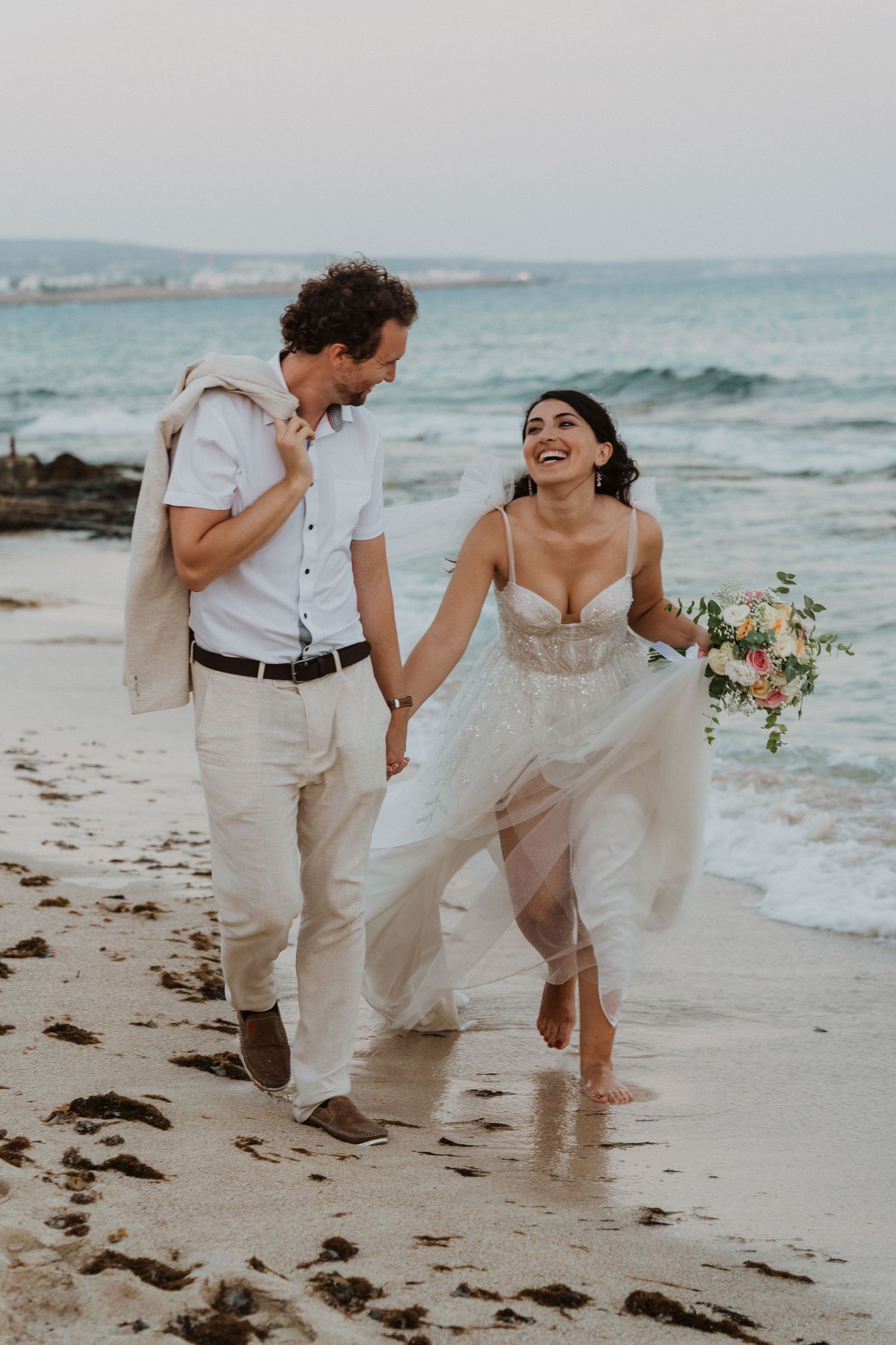 Happy newlyweds running and laughing together on sandy beach at sunset.