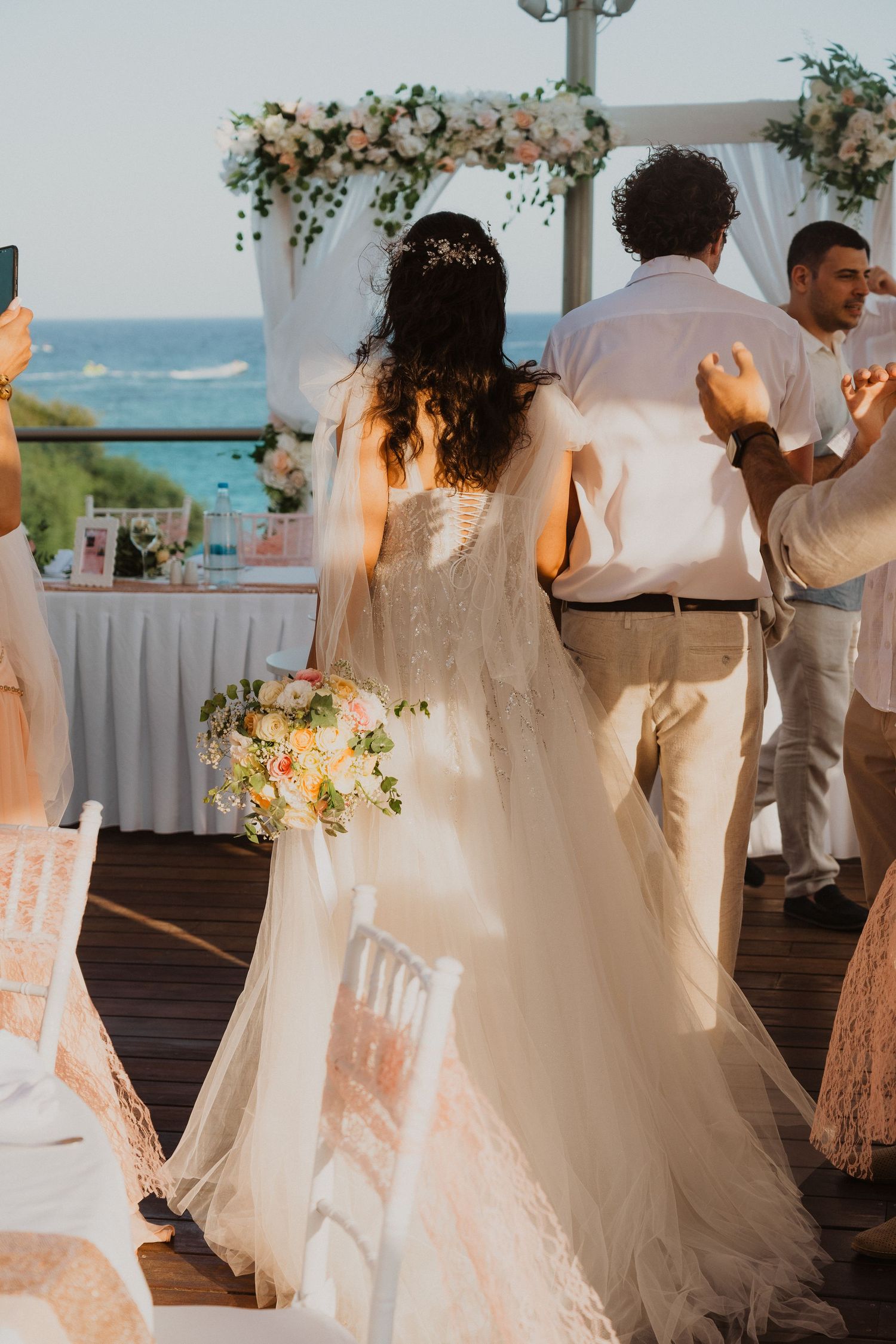 A bride in a flowing white gown stands at an oceanside reception decorated with floral arrangements.