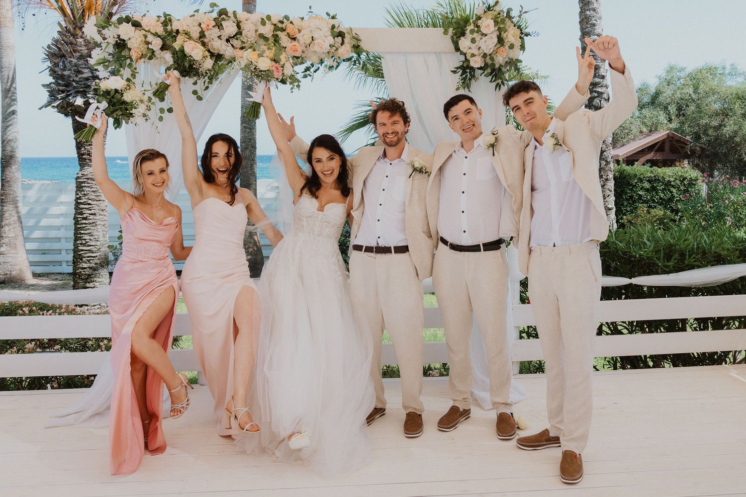 Wedding party in light summer attire poses together under decorated arch by the ocean.