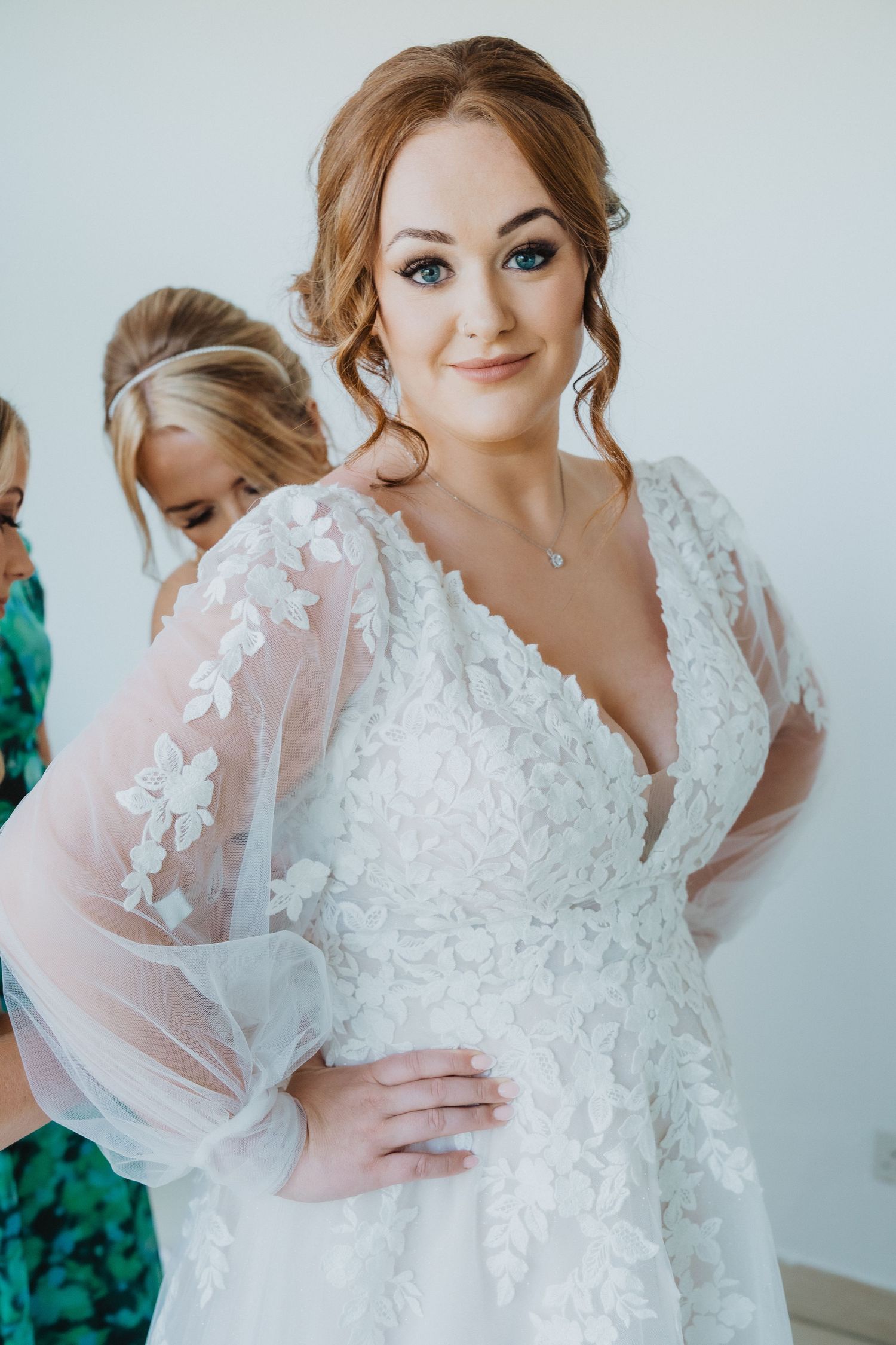 A bride in a stunning white lace wedding gown with sheer sleeves poses during her wedding preparations.