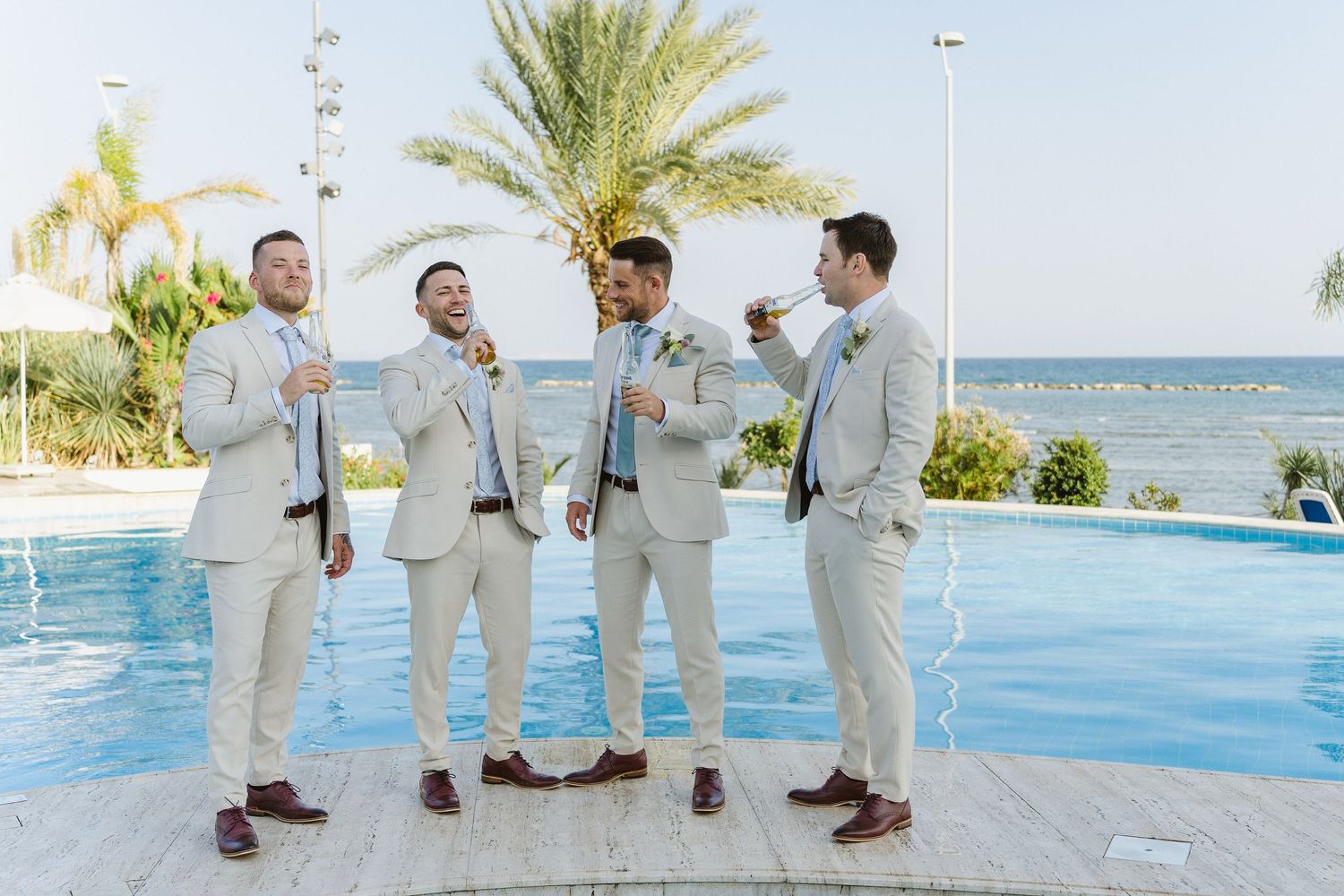 Wedding party in light suits enjoying drinks by poolside with ocean view.