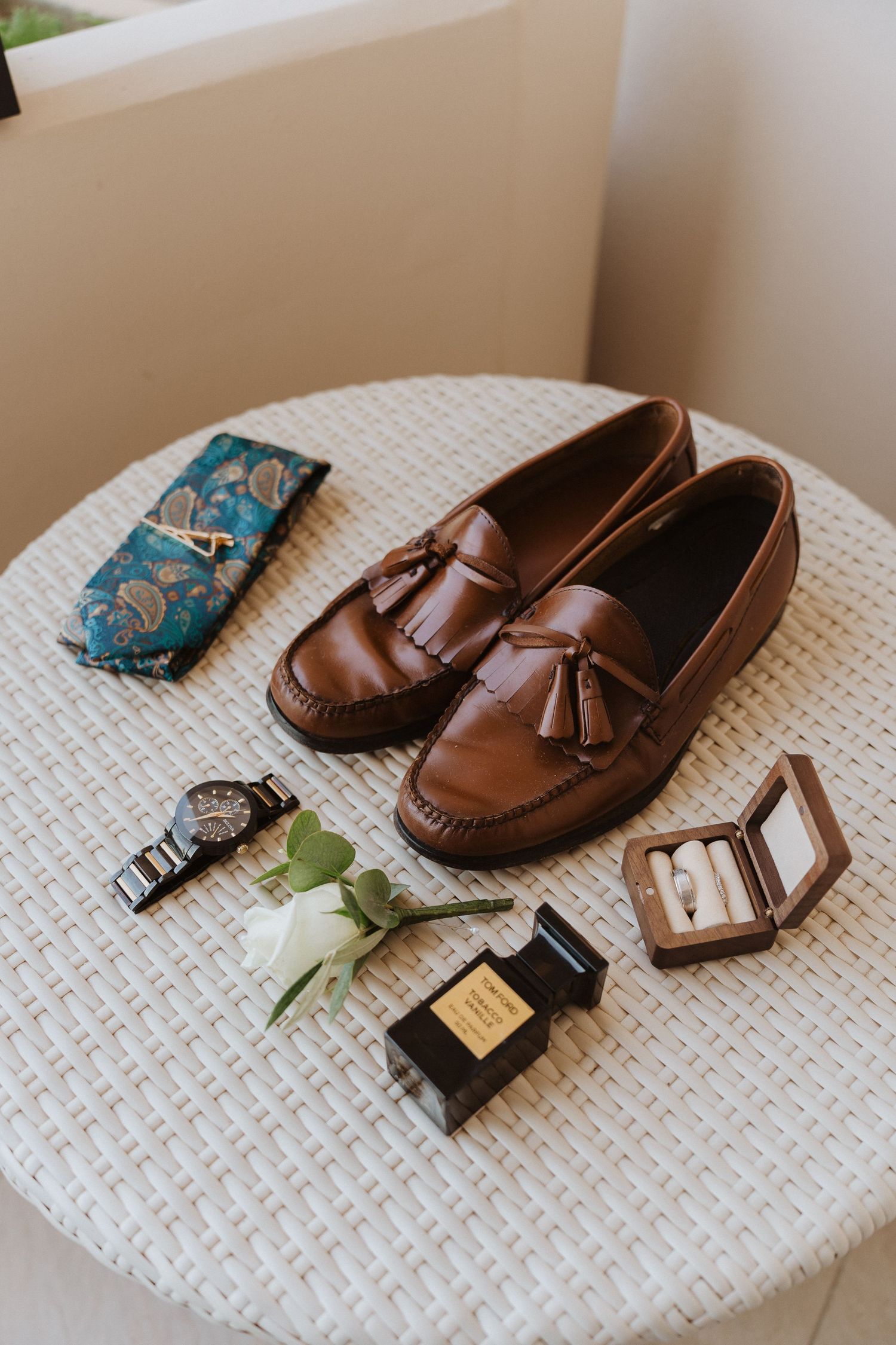 Brown leather loafers with tassels displayed alongside a watch, perfume bottle and floral accessories.