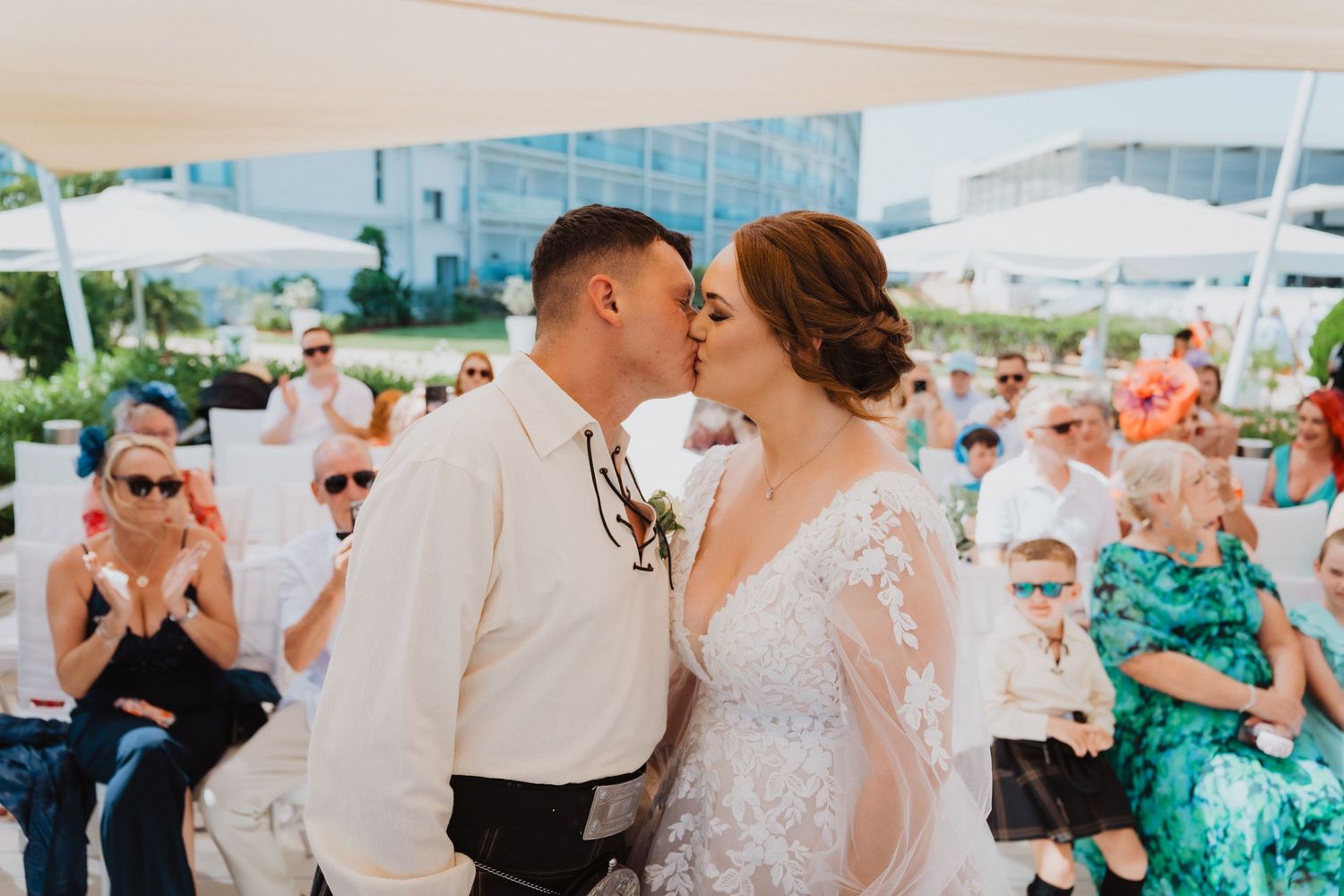 A tender wedding kiss captured at a beachfront ceremony with guests watching.