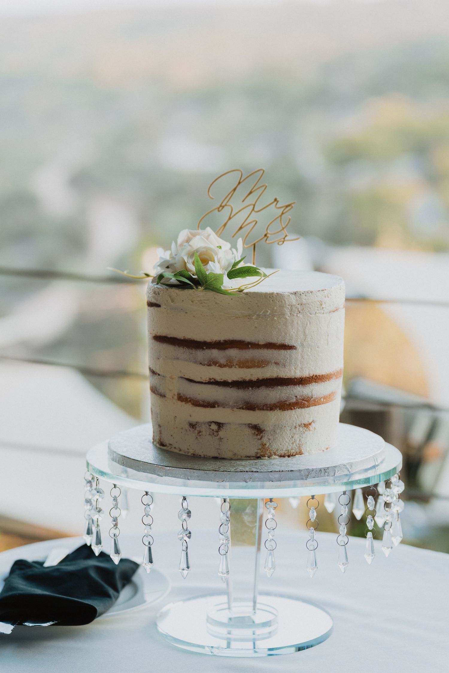 A minimalist semi-naked wedding cake with white flowers on top displayed on a crystal cake stand.