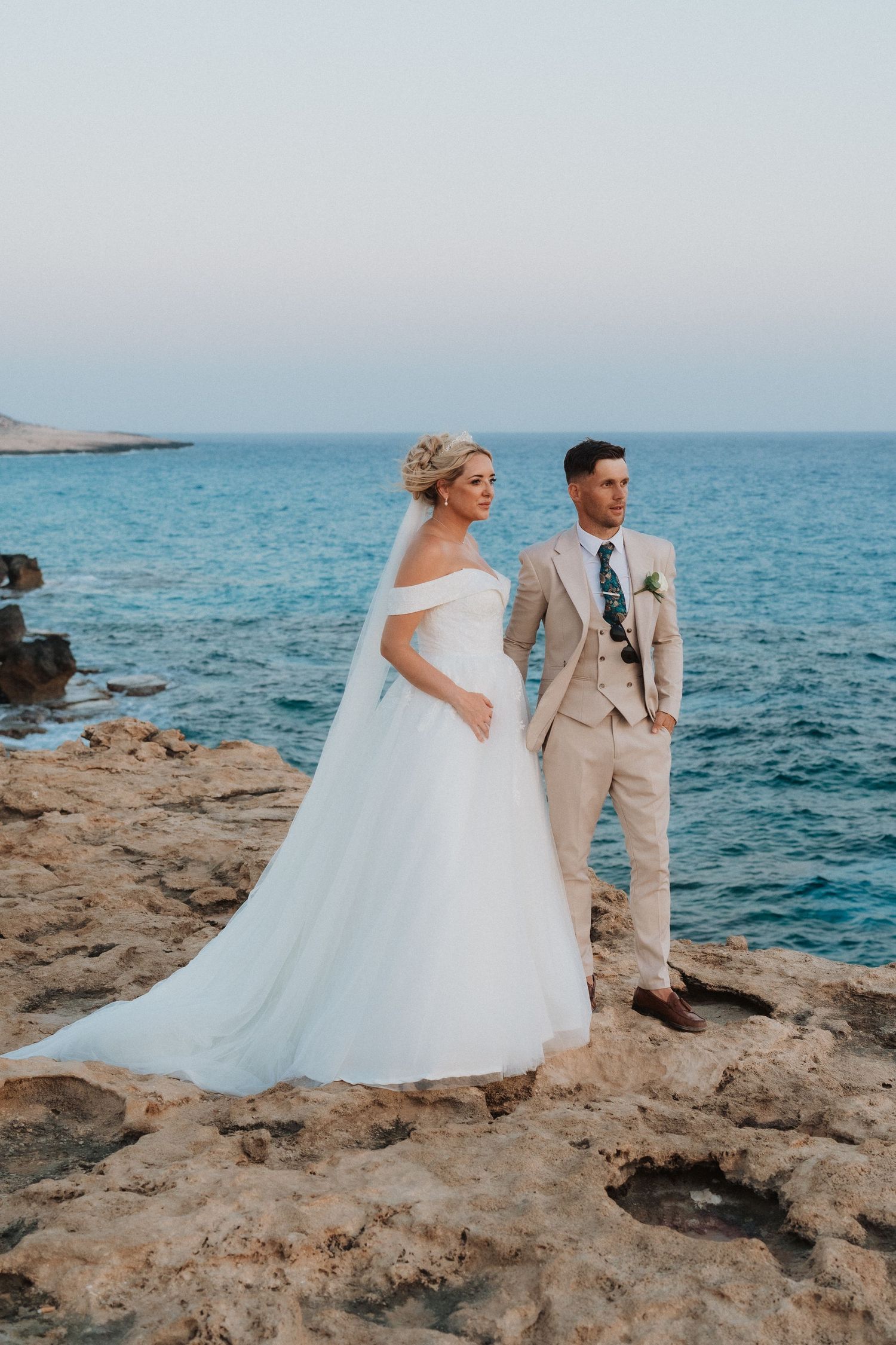 Wedding couple standing on rocky coastal cliffs overlooking bright blue Mediterranean waters.