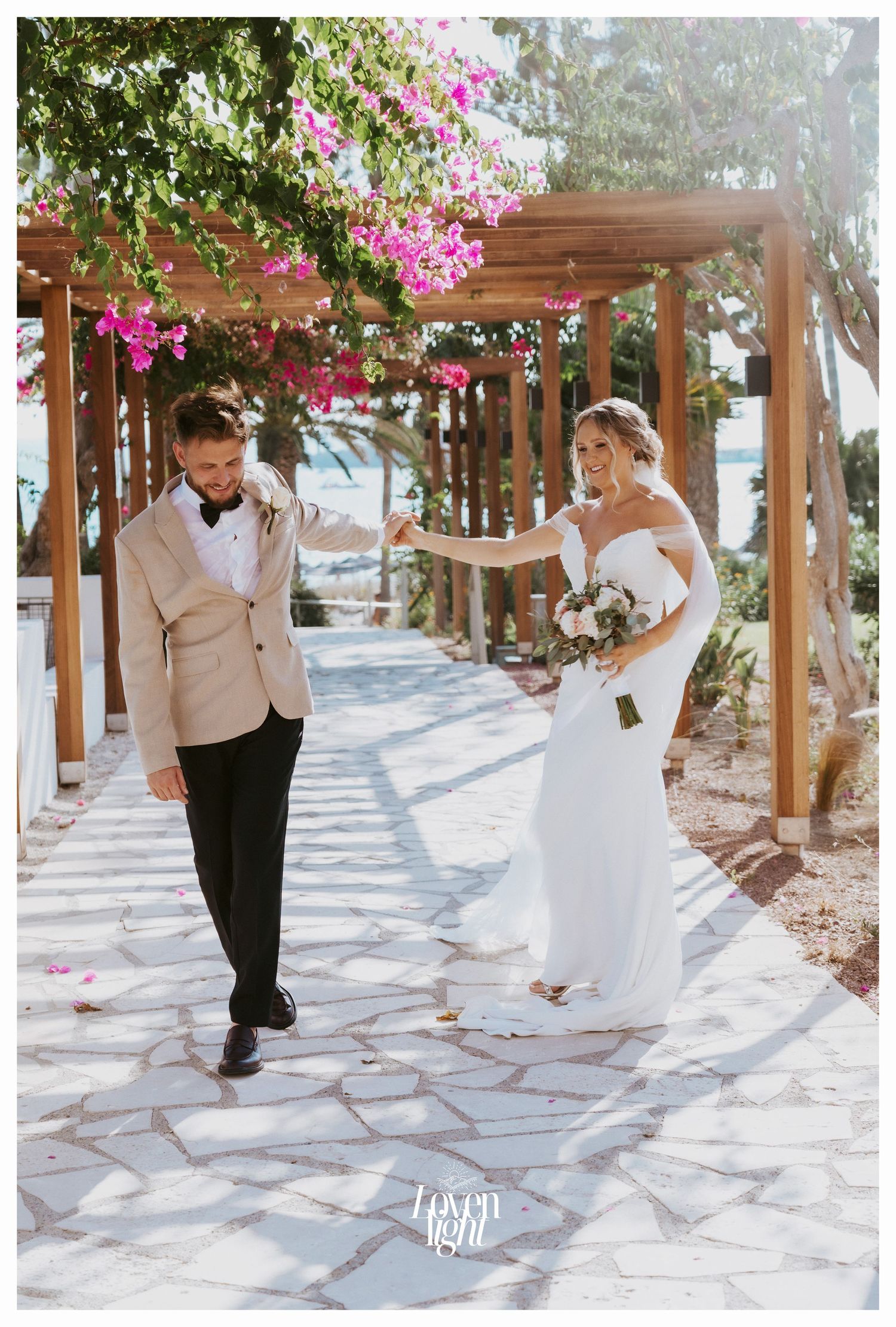 Playful wedding couple walking under a pergola decorated with bright pink flowers.