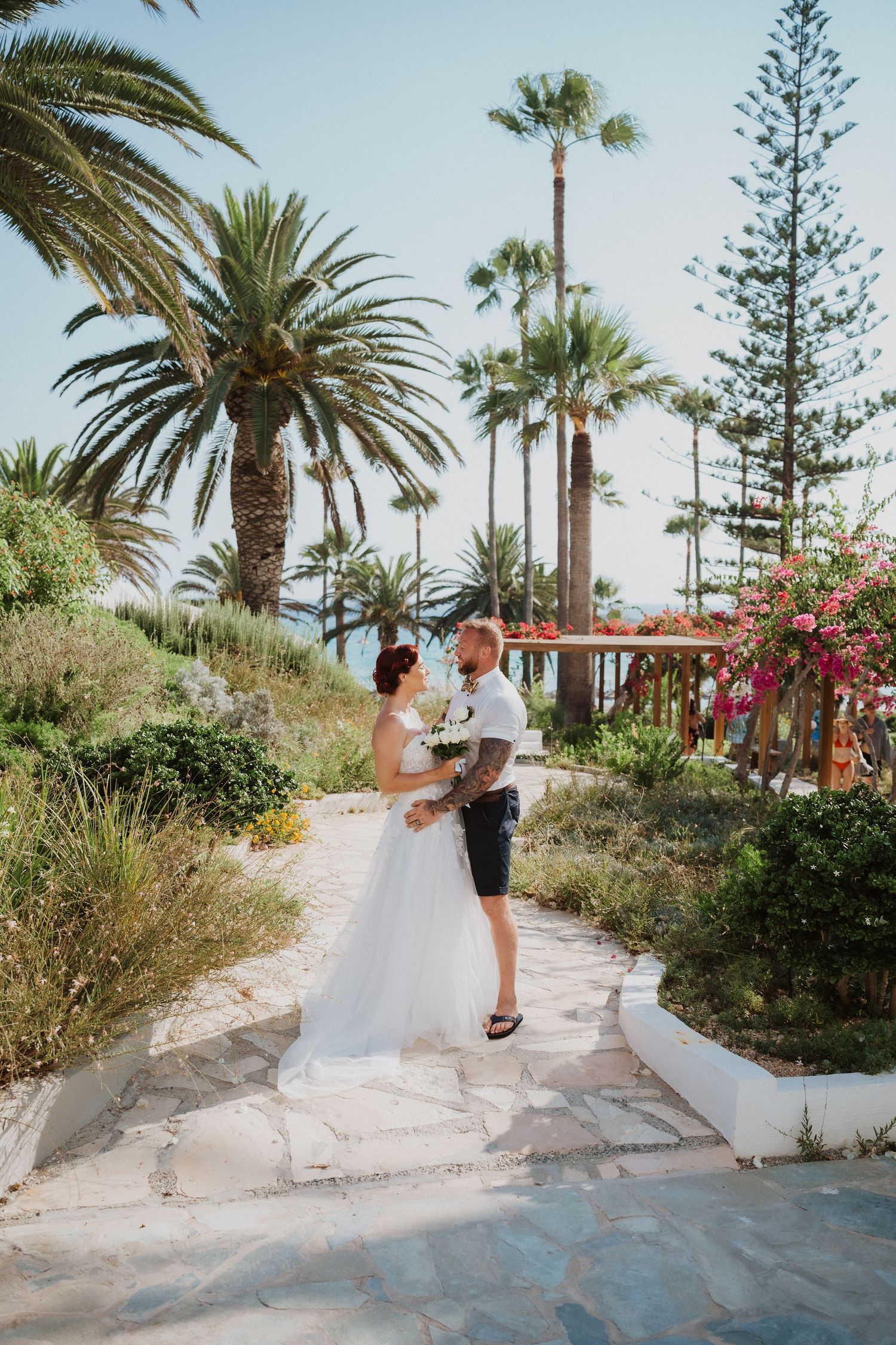 A wedding couple stands together on a Mediterranean path surrounded by palm trees and coastal views.
