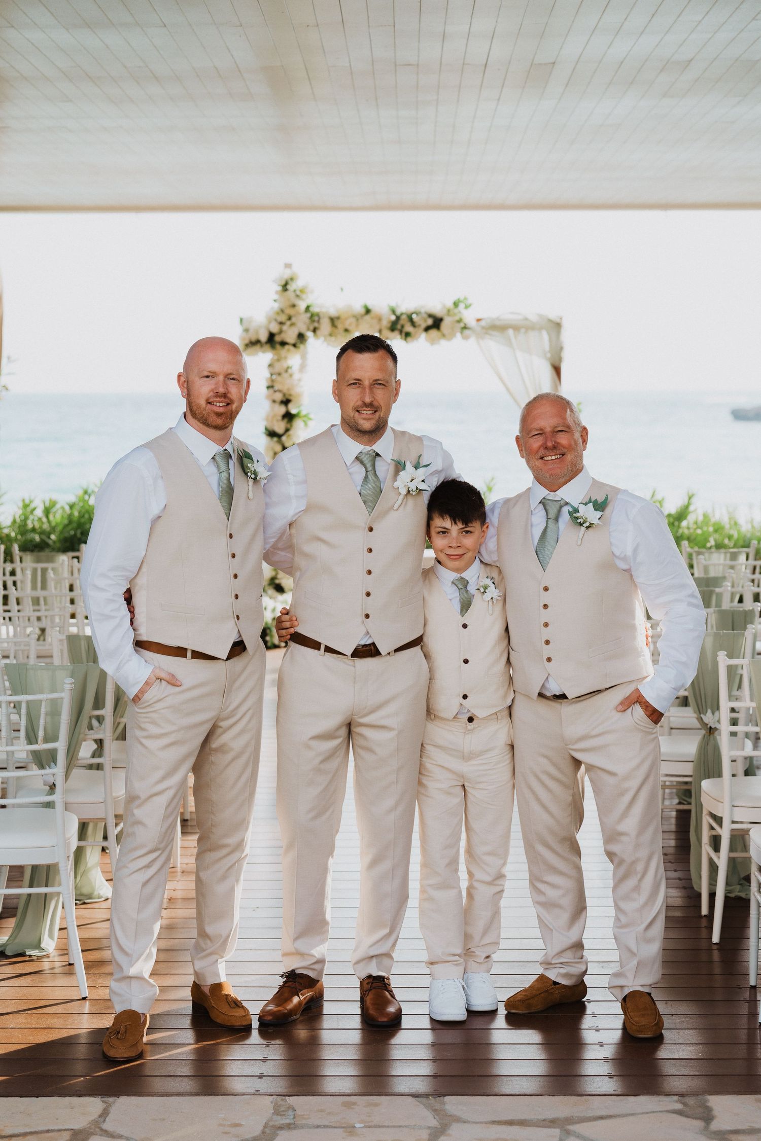 A wedding party in matching cream vests and slacks poses together on a covered oceanfront terrace with floral decor.