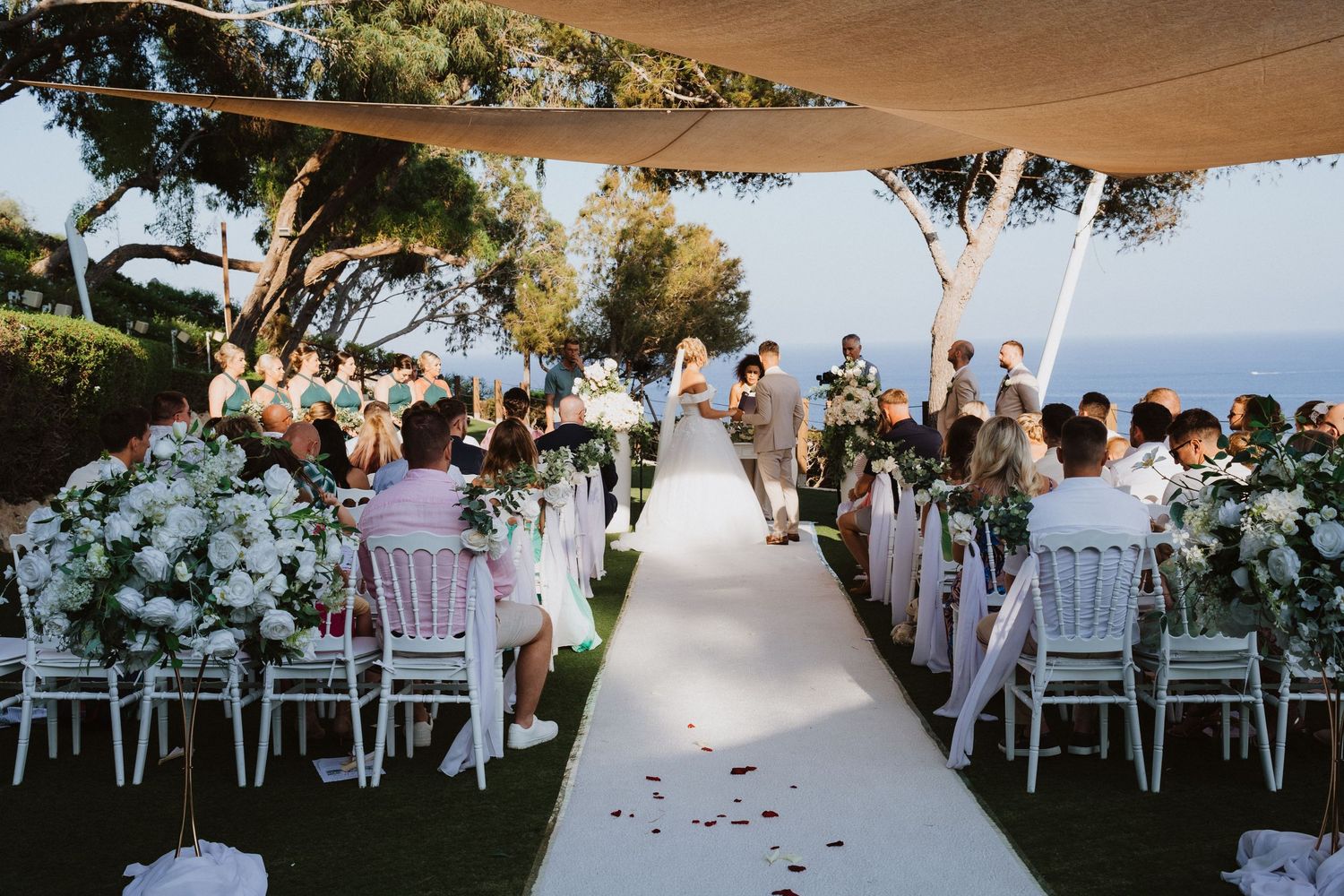 Outdoor wedding ceremony setup with white chairs and floral decorations overlooking the ocean under a canopy.