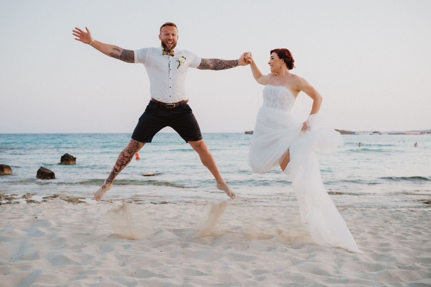 Wedding couple leaping joyfully together on a beach with ocean waves in background.