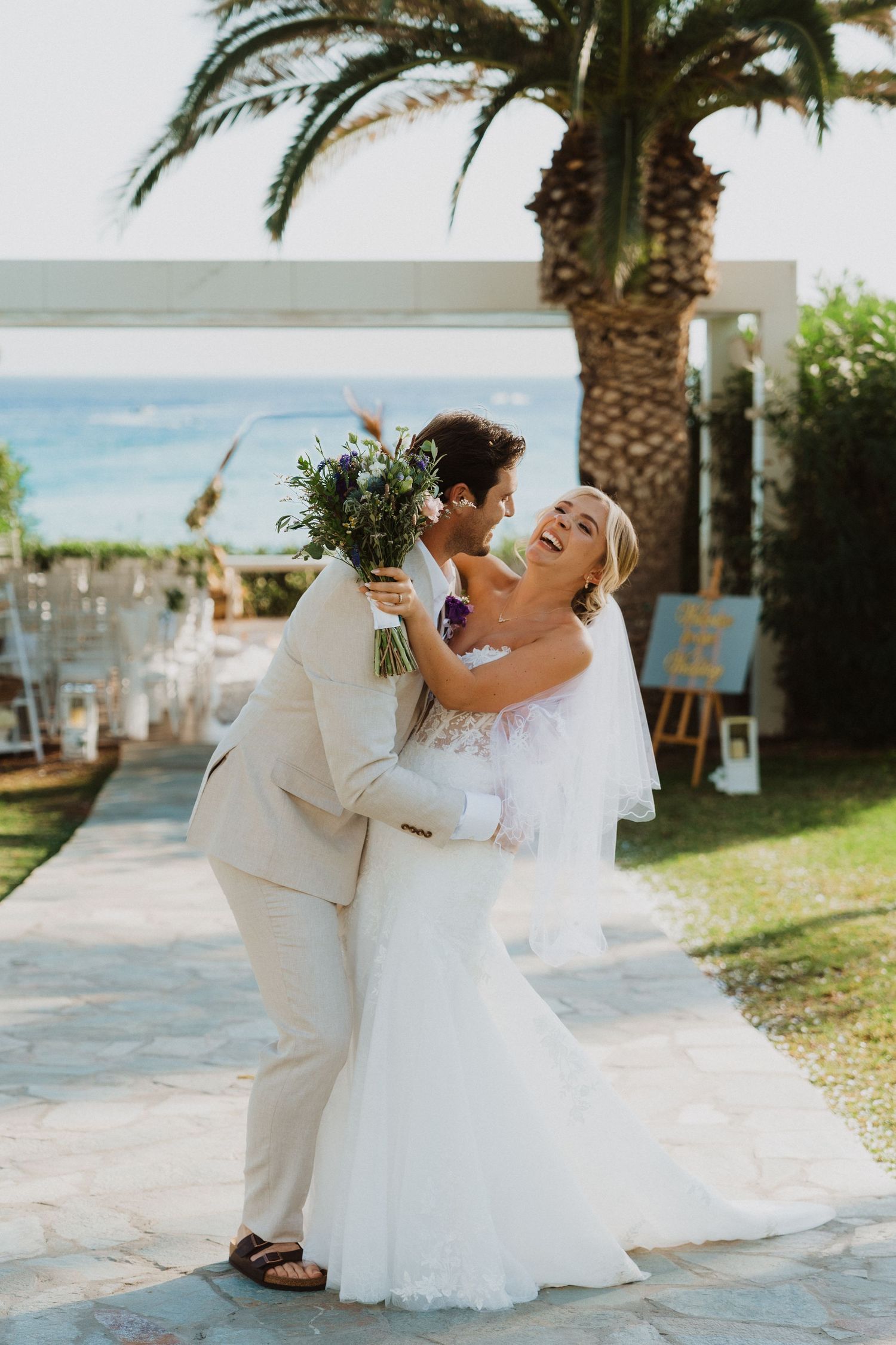 A joyful wedding couple dances together on a stone pathway near palm trees.