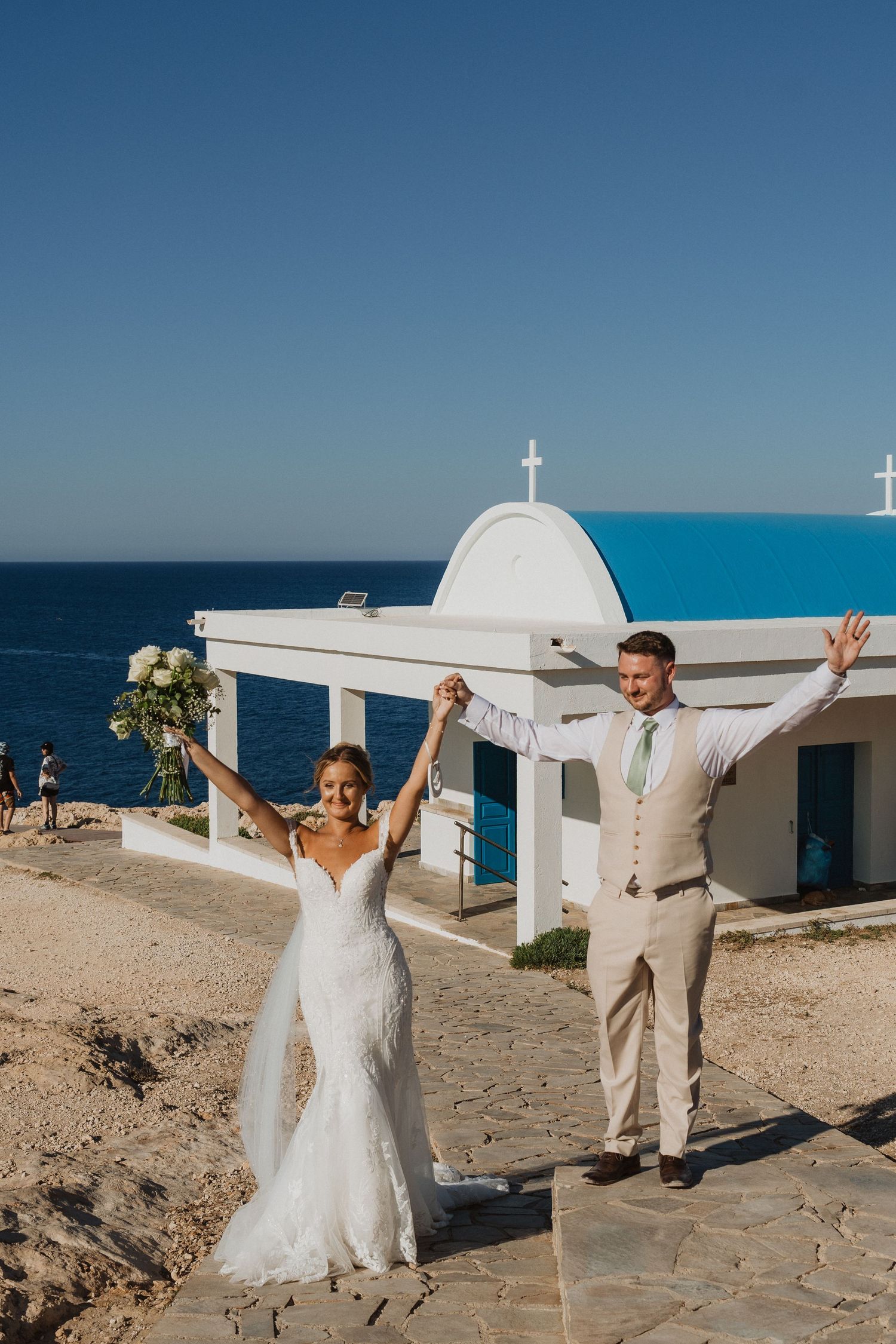 A newlywed couple celebrates joyfully outside a white Mediterranean church overlooking the bright blue ocean on their wedding day.