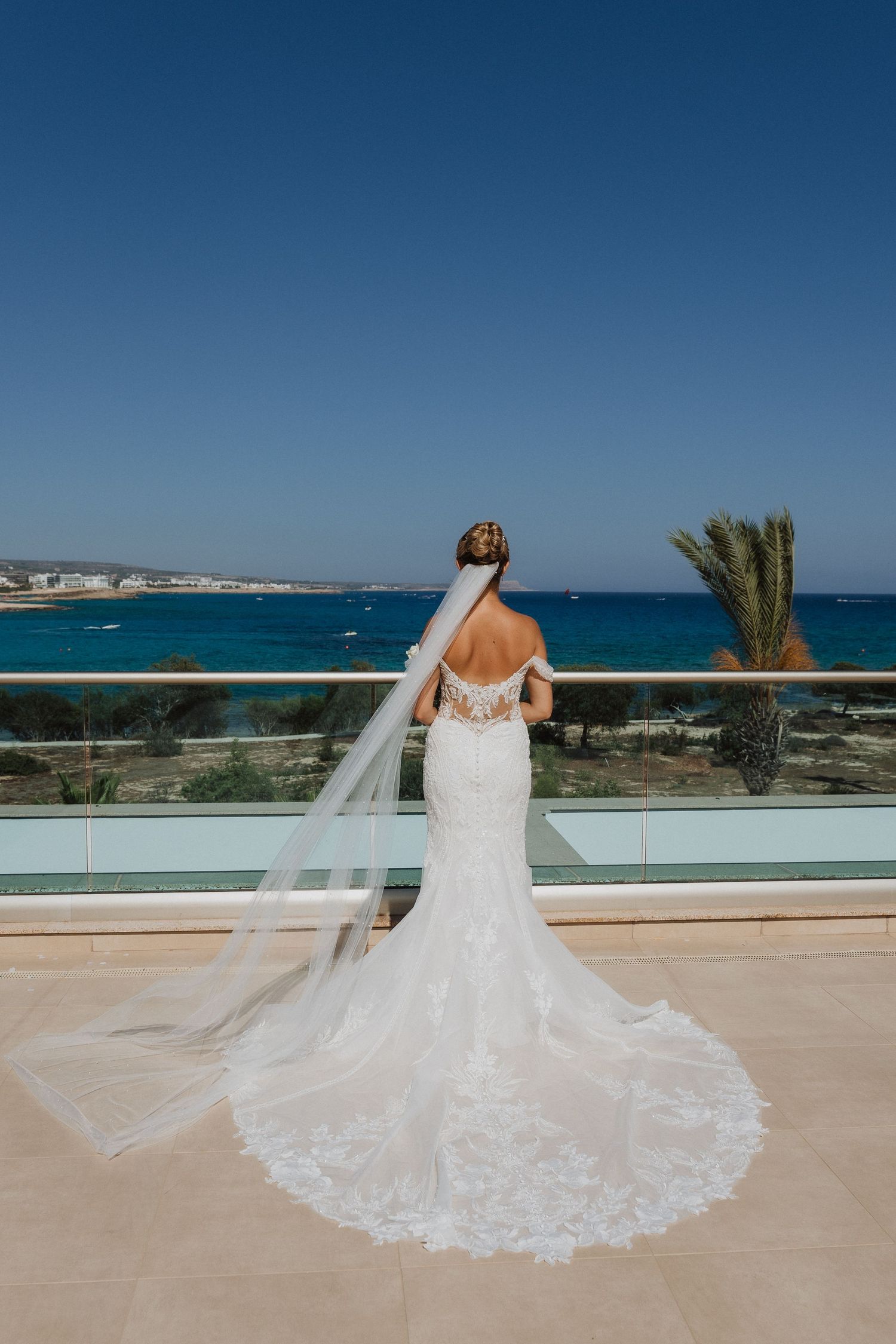 A bride in a lace mermaid wedding dress and veil stands on a balcony overlooking a beautiful Mediterranean coastline.
