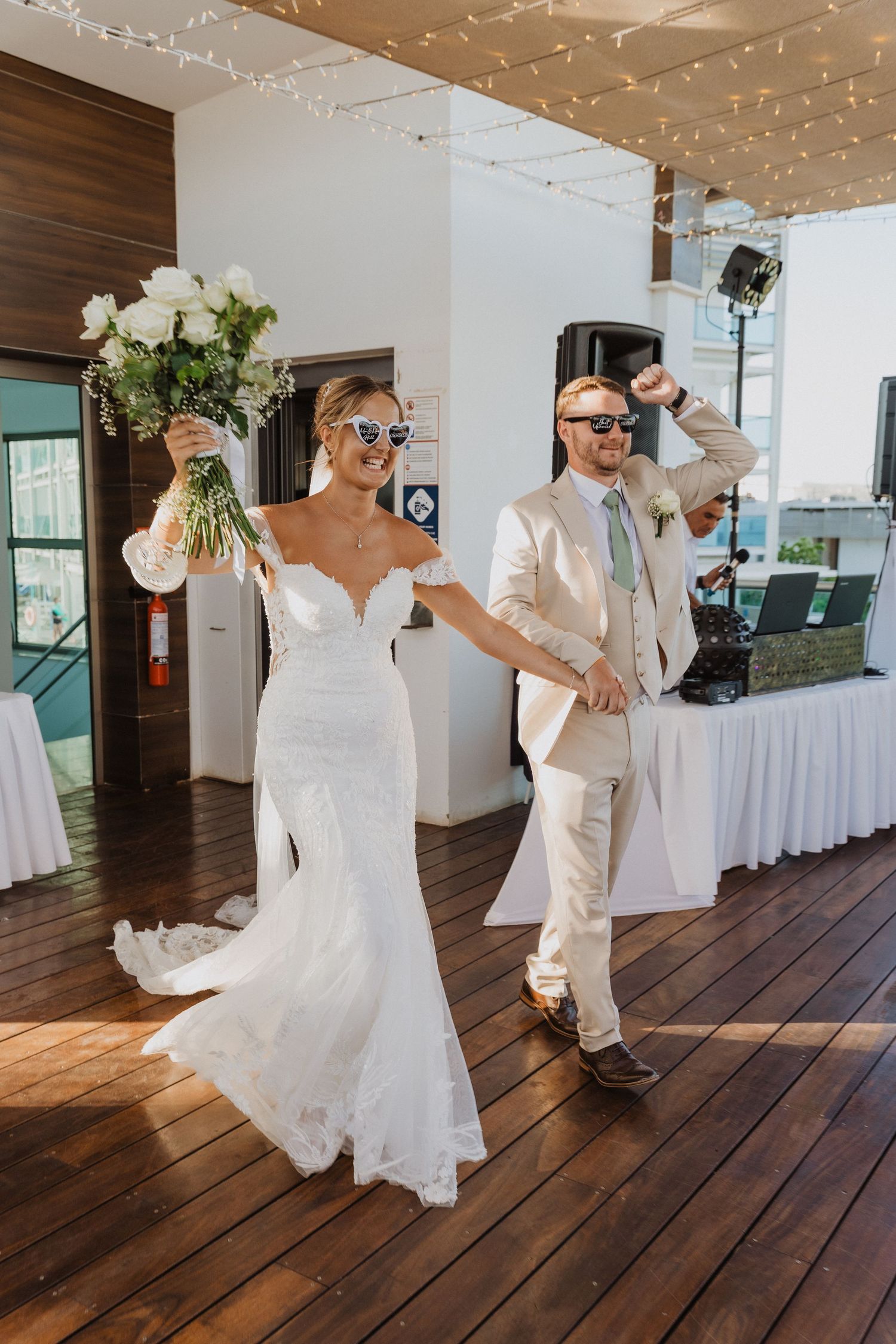 A couple in white attire dances together on a wooden floor during what appears to be a wedding celebration.
