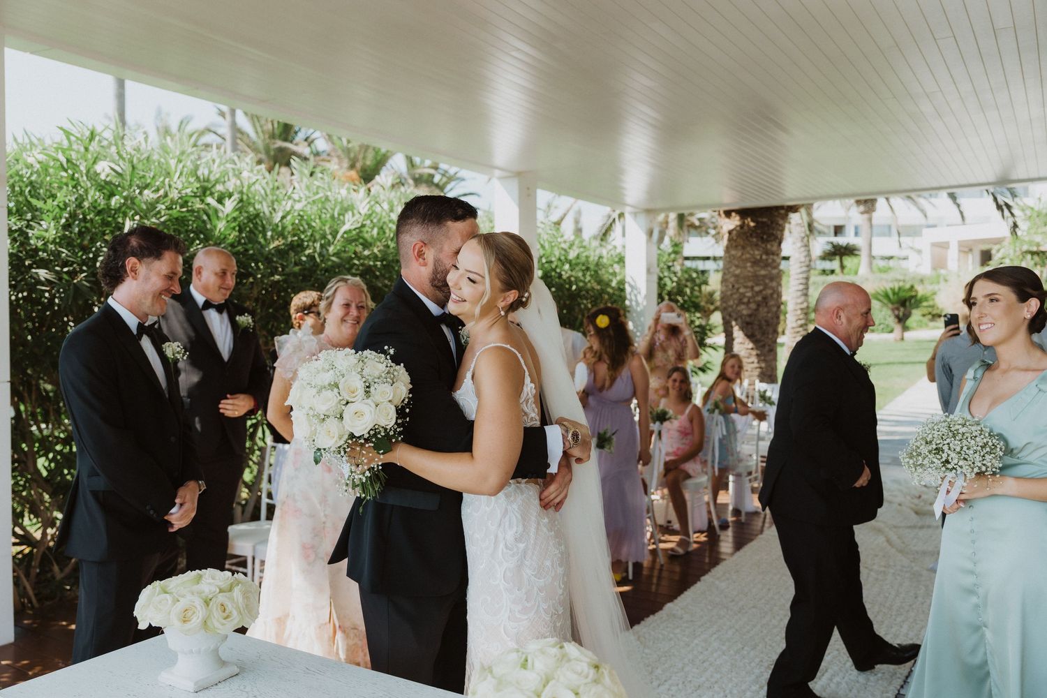 Wedding guests dancing and celebrating at an elegant indoor reception venue with white decor and palm trees.