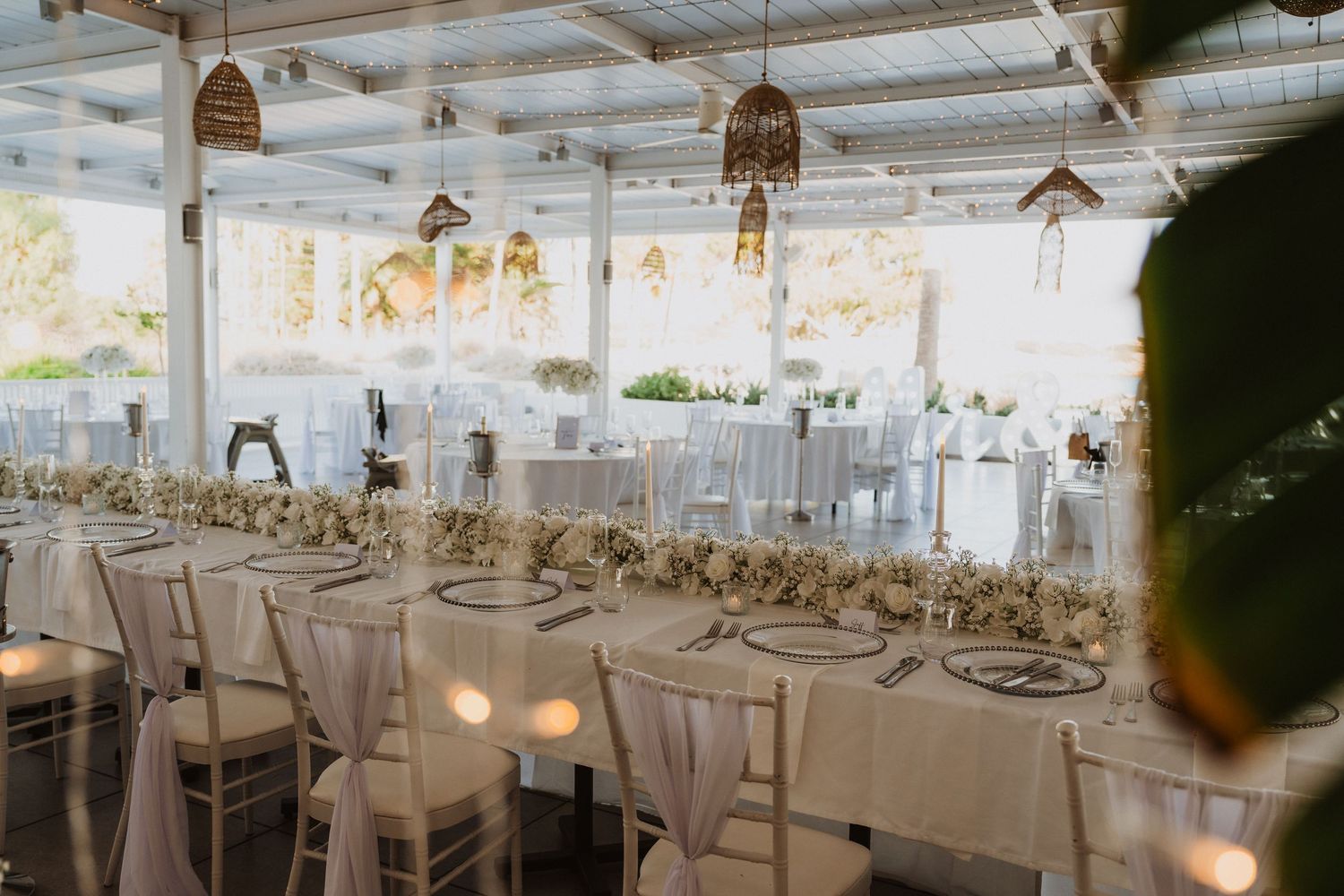 Light-filled wedding reception venue with white tablecloths and hanging wicker pendant lights.