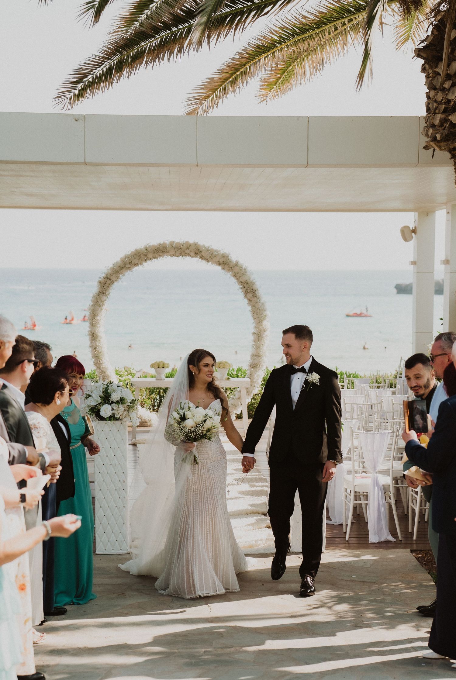 Bride and groom walk down aisle at beachfront wedding ceremony with floral arch and ocean backdrop.