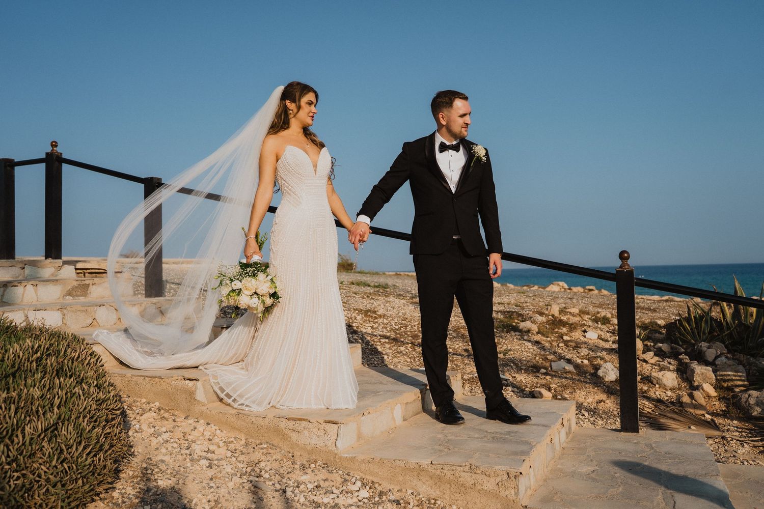 Bride and groom hold hands on rocky coastal steps with a stunning blue ocean backdrop.