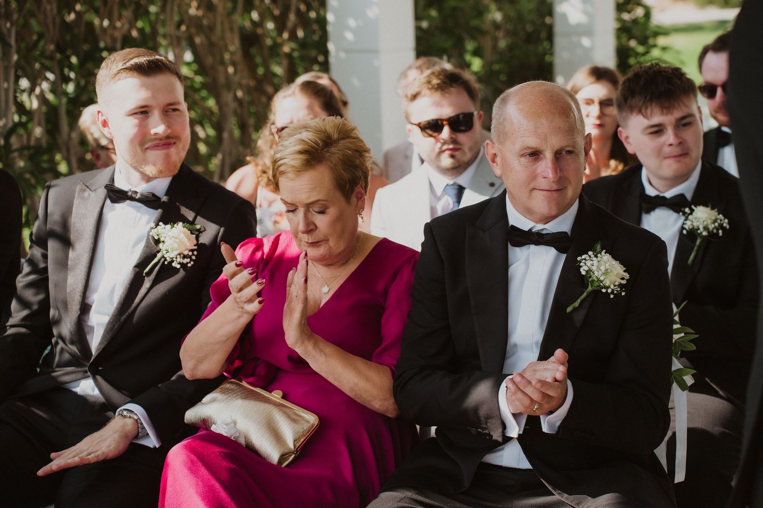 Emotional wedding guests in formal attire applauding during an outdoor wedding ceremony.