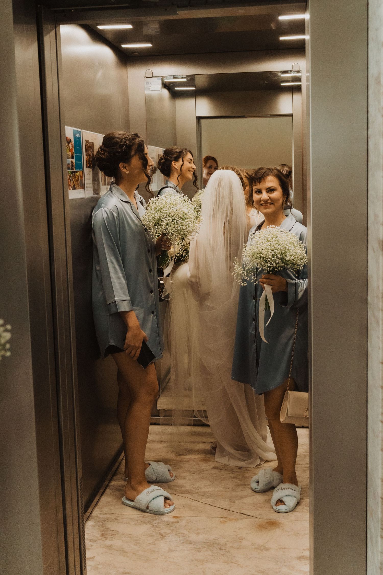 Bridesmaids in blue robes holding baby's breath bouquets with bride in white gown and veil inside elevator.