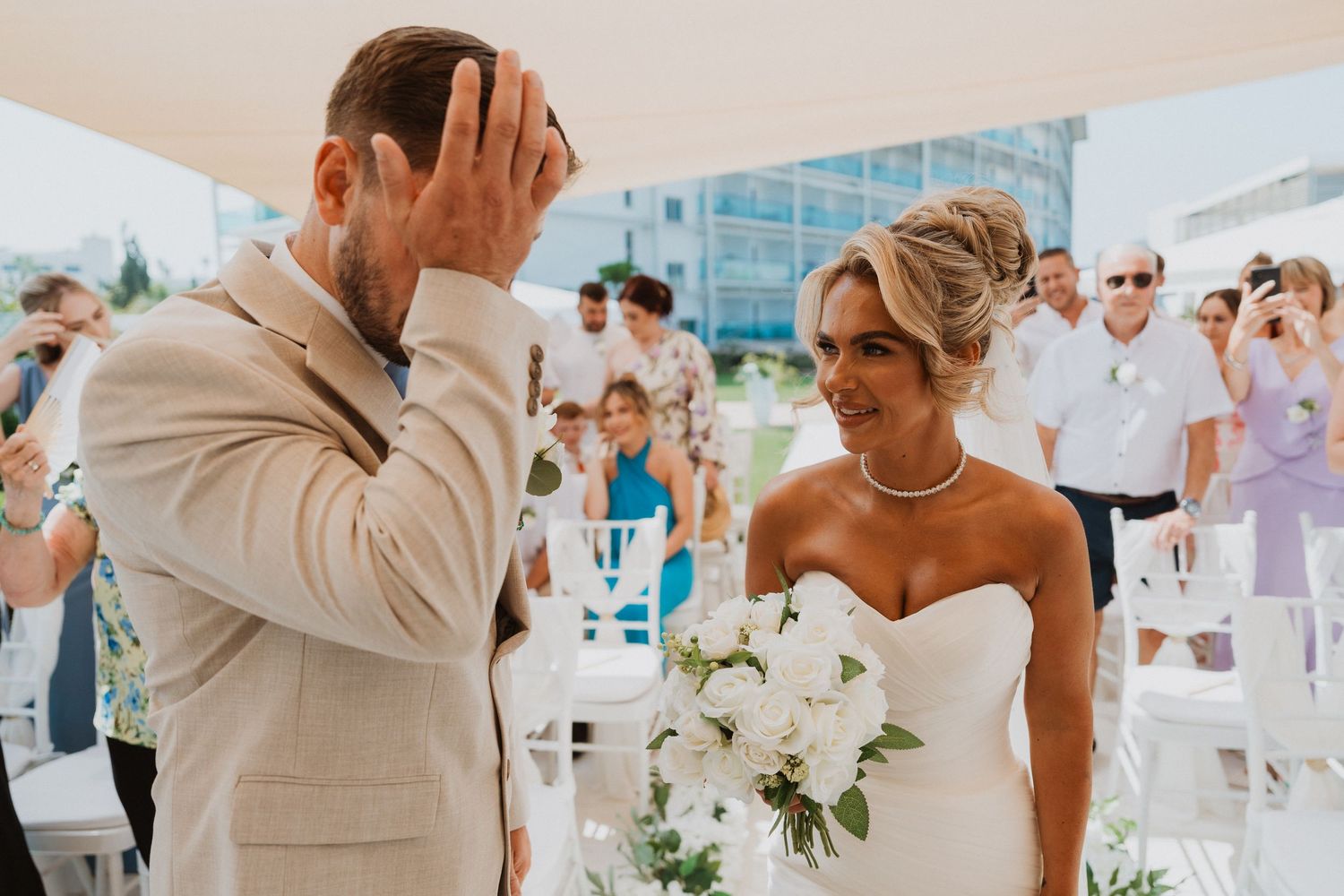 Emotional groom covering face as bride smiles holding white bouquet at outdoor wedding ceremony with guests.