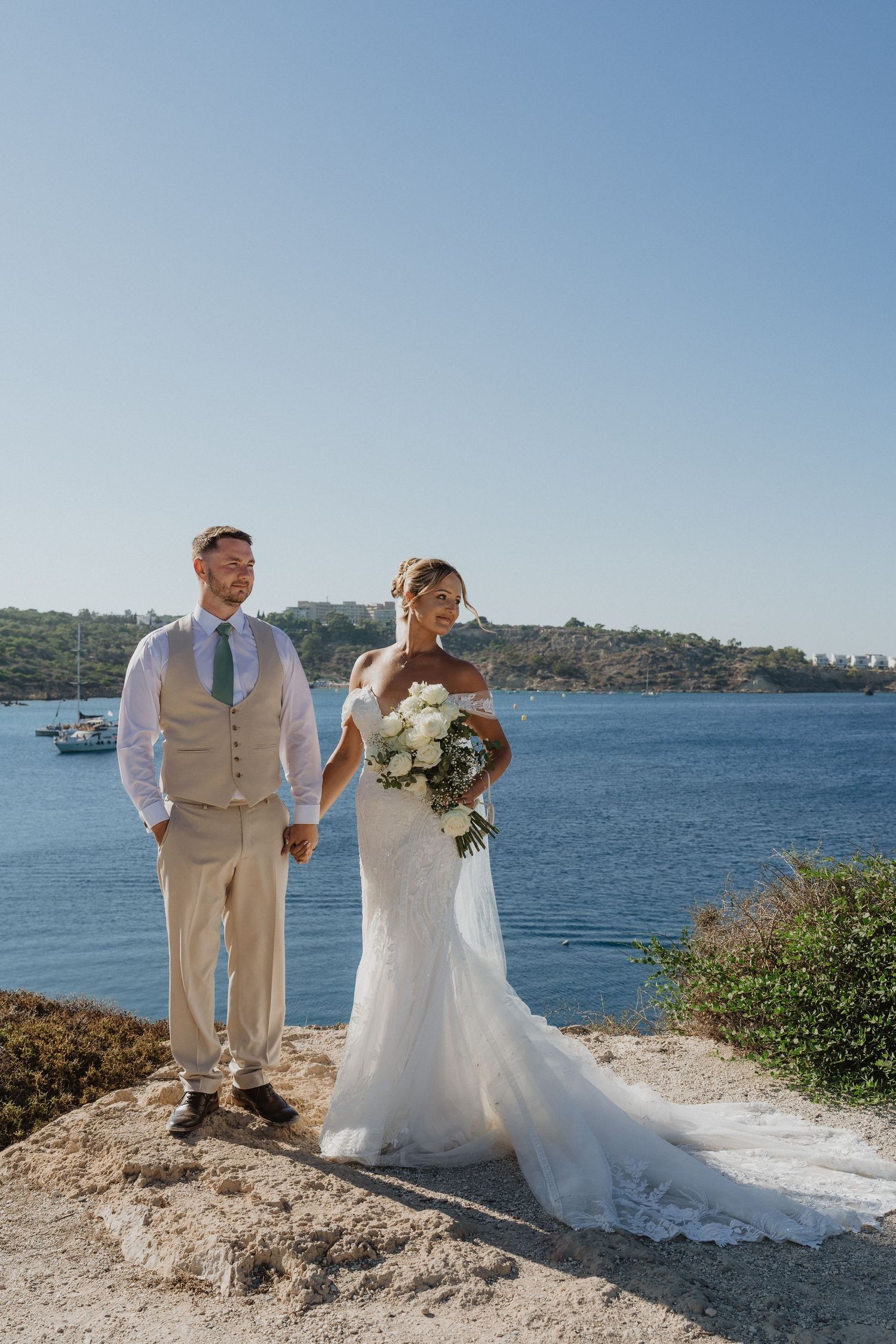 A couple in wedding attire poses along a scenic waterfront with blue ocean views and green hillsides in the background.