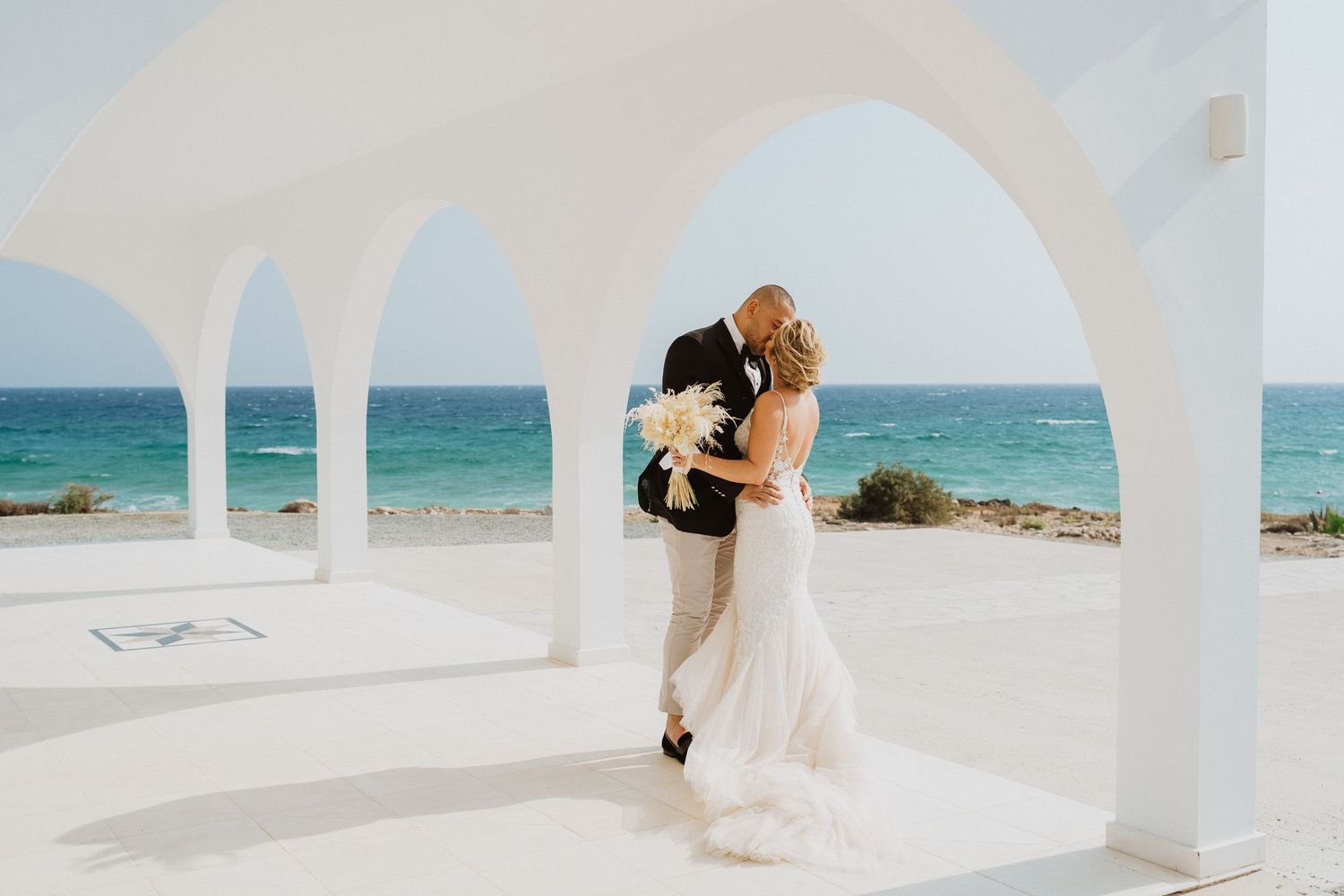 A romantic beach wedding ceremony with a couple embracing under a white arch by turquoise ocean waters.