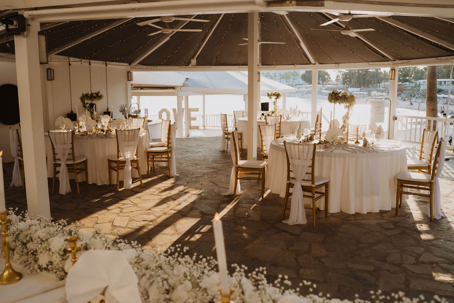 Elegant outdoor wedding reception under a pavilion with gold chairs, white tablecloths, and floral centerpieces by the water.