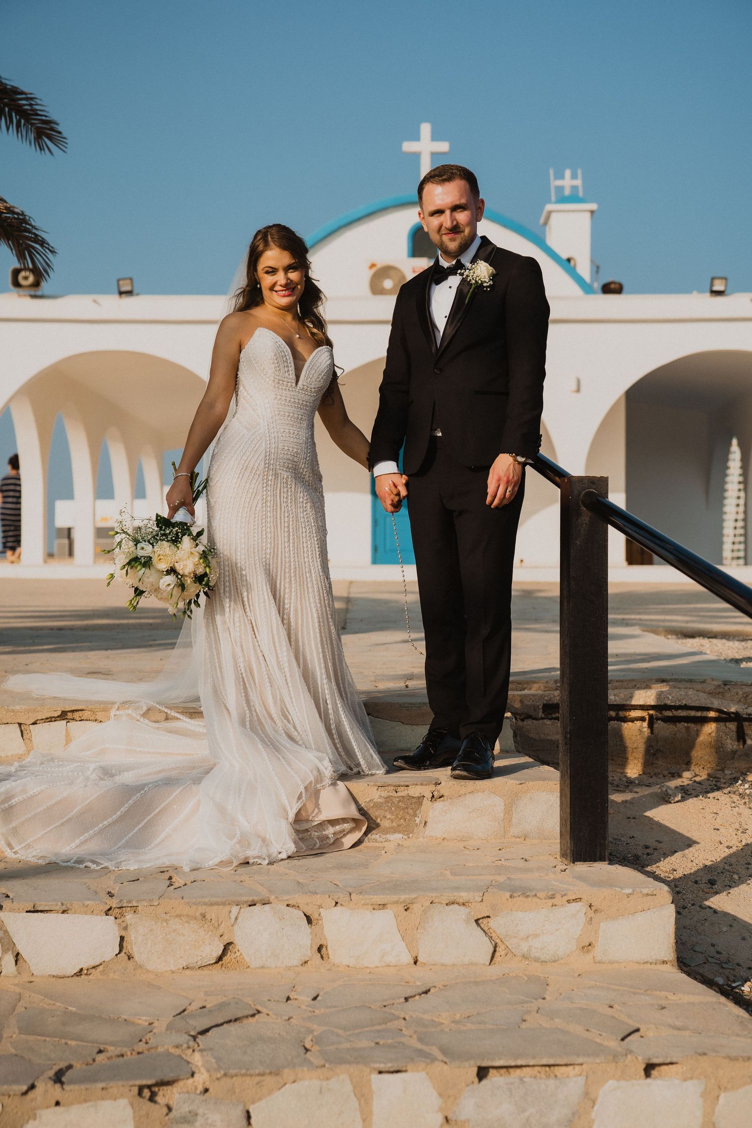 Bride and groom pose on stone steps before a white Greek Orthodox chapel under a clear blue sky.