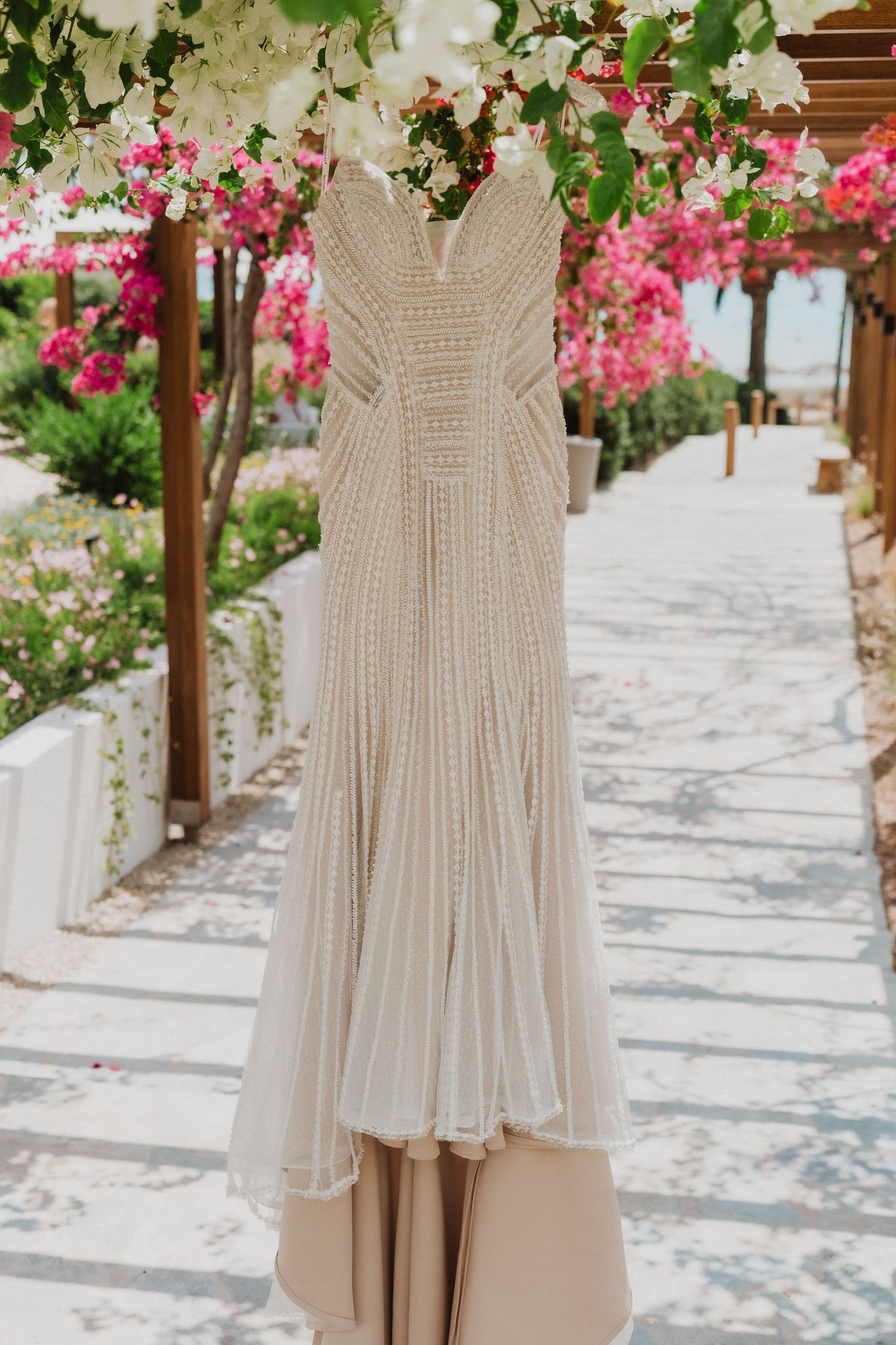 Elegant beaded ivory wedding gown hanging outdoors under pink bougainvillea flowers at a garden venue.