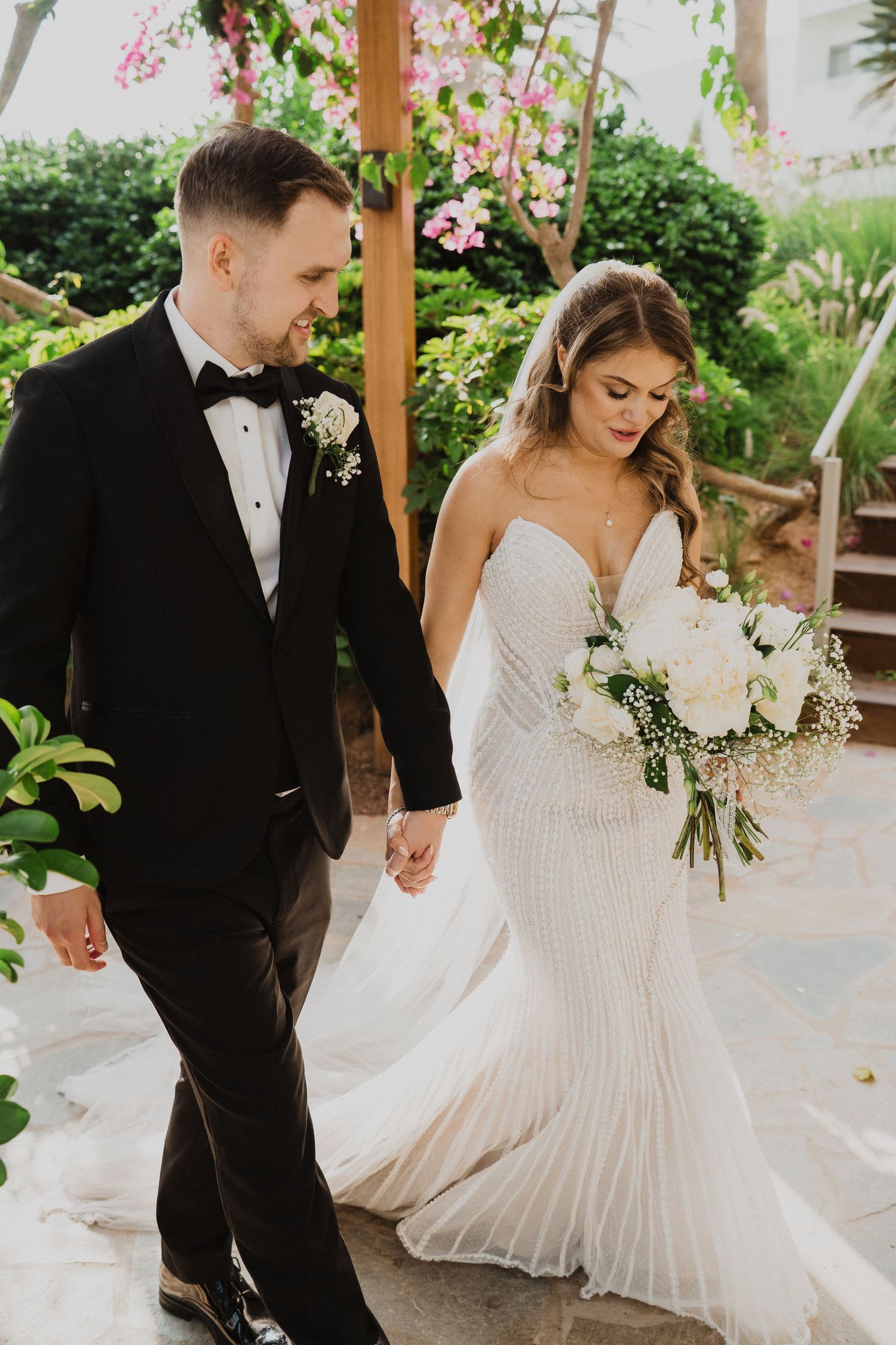 Bride and groom hold hands walking through lush garden, bride carrying white bouquet in beaded gown.