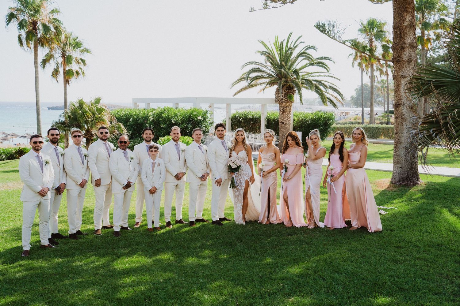 Wedding party members in white and blush pink attire gather on grass with palm trees in background.