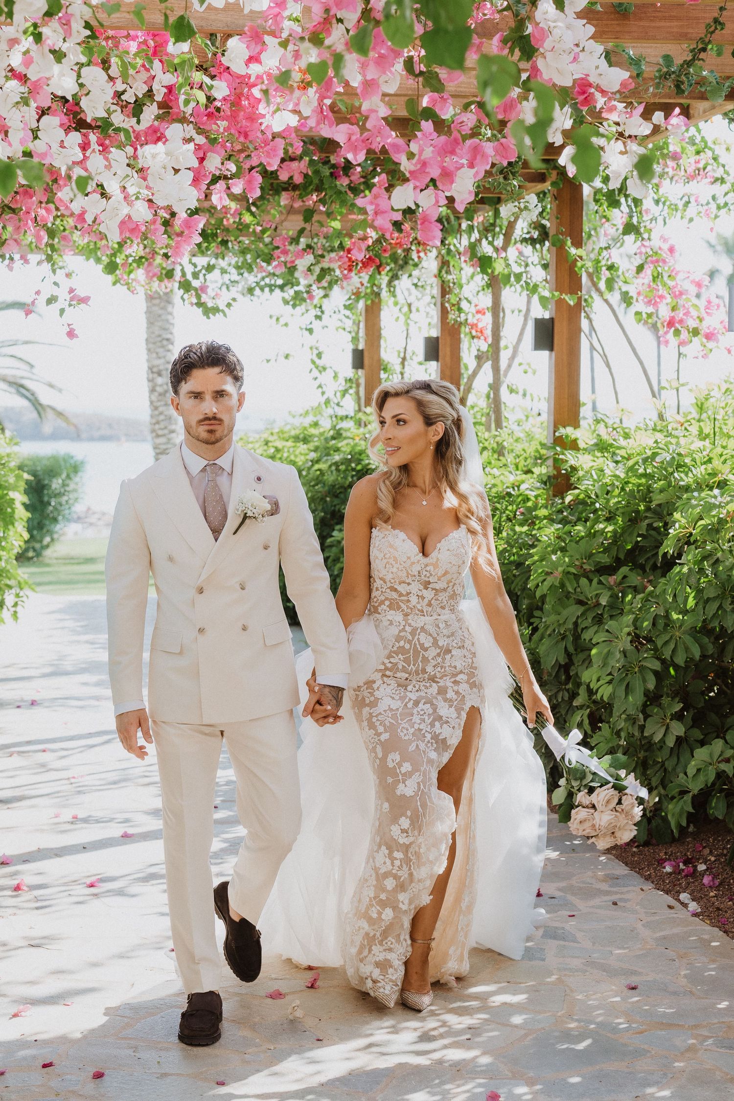 A couple walks beneath a romantic pergola covered in pink and white flowering vines at an elegant outdoor venue.