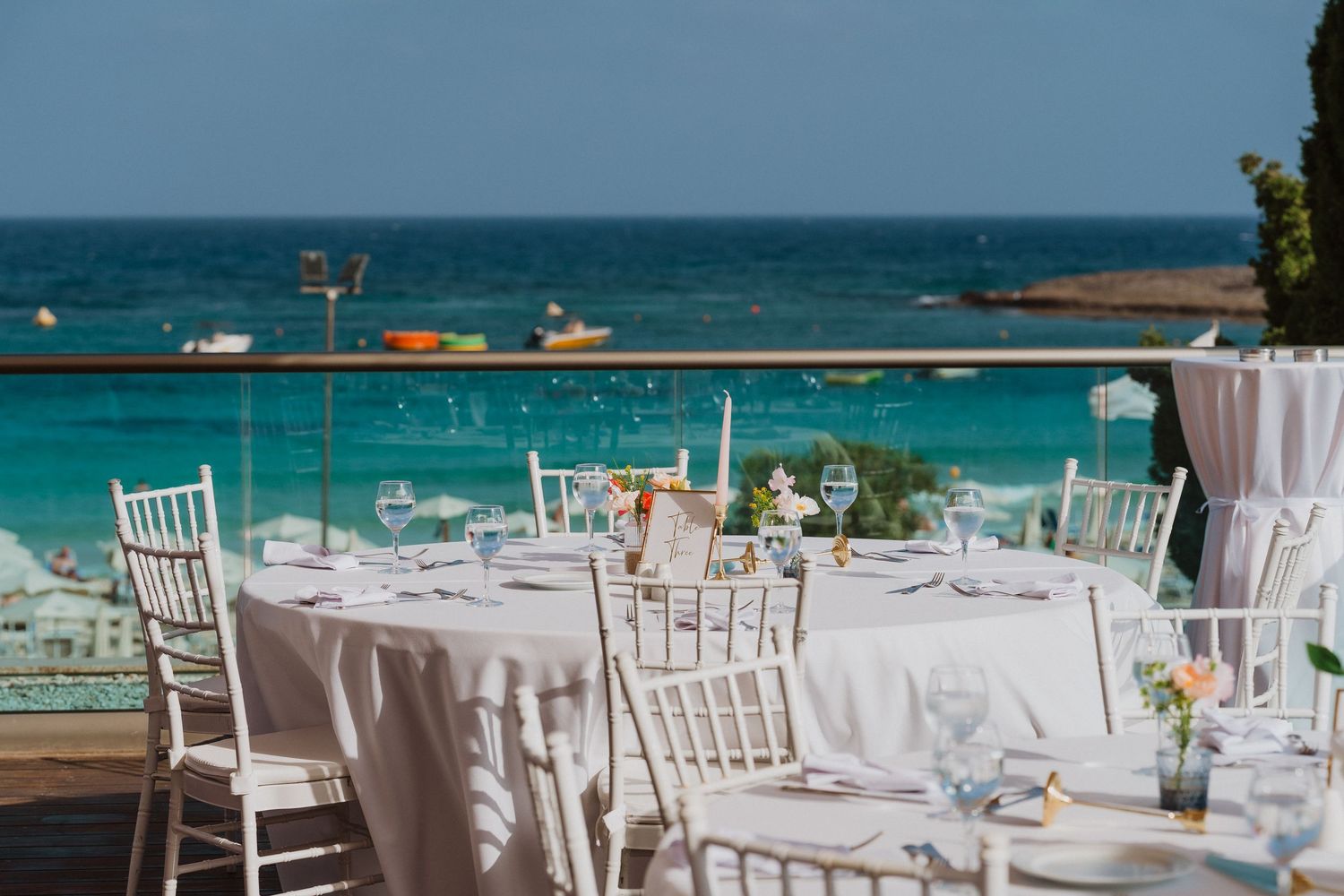 White dining tables and chairs set up for a beachfront wedding reception with turquoise ocean views in the background.