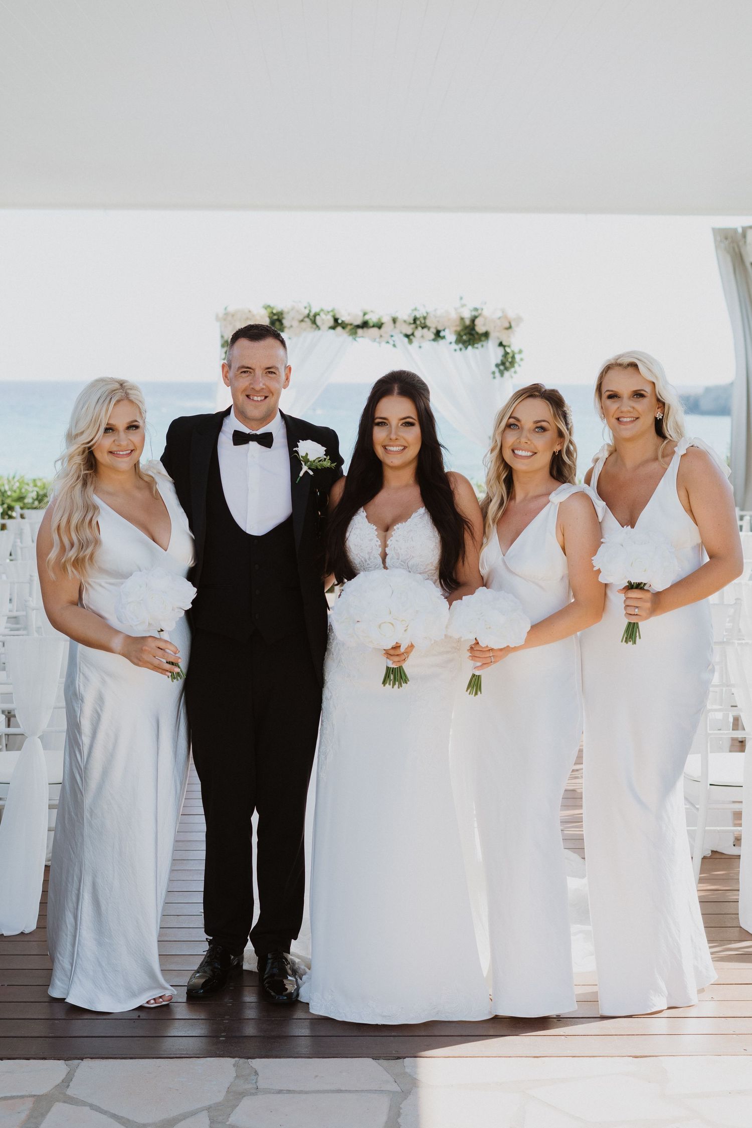 Wedding party in white dresses and black tuxedos poses together at an elegant beachside venue.