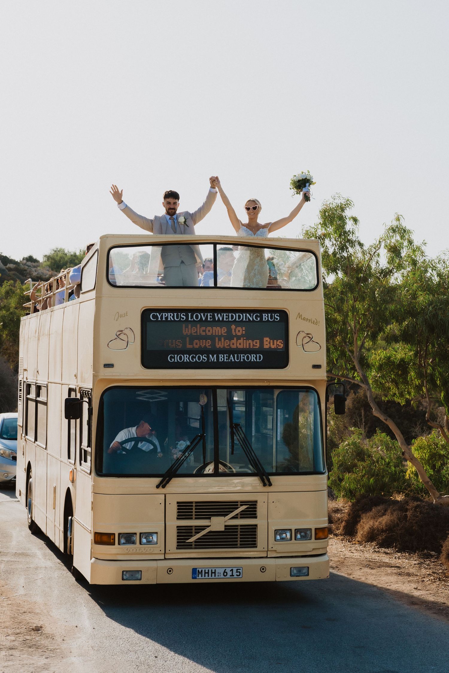 A cream-colored double-decker wedding bus with celebrating passengers on a coastal road.