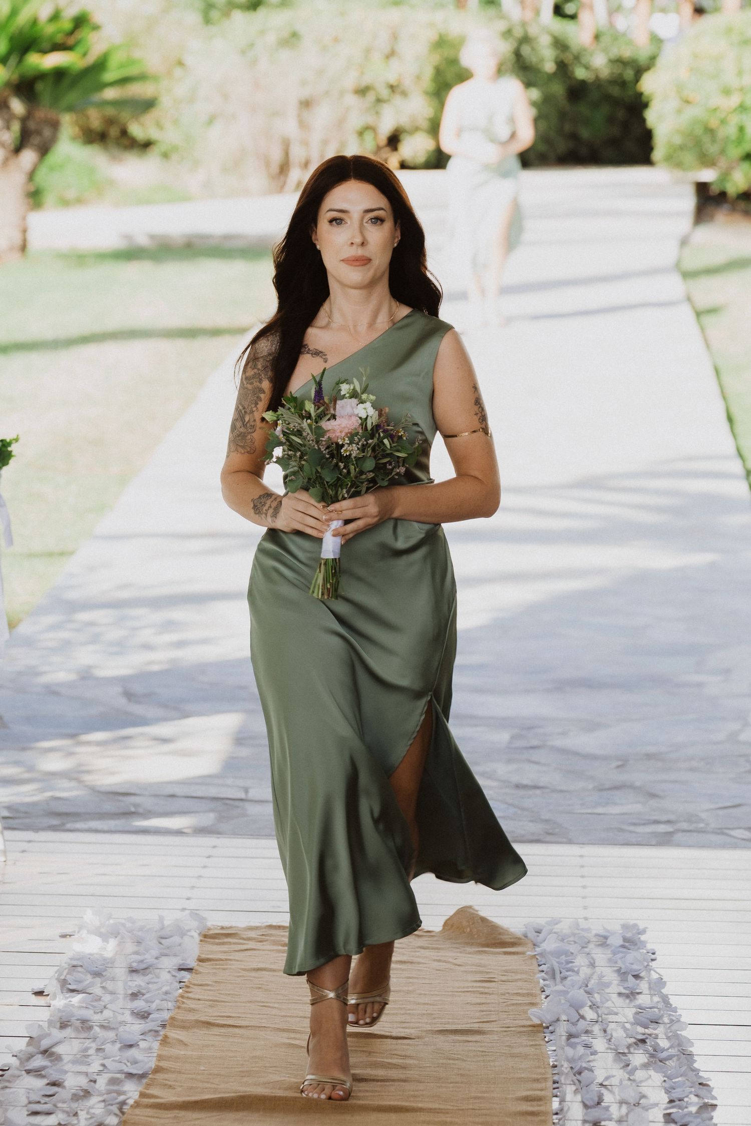 Bridesmaids in sage green dresses walk down a petal-covered aisle at an outdoor wedding venue.