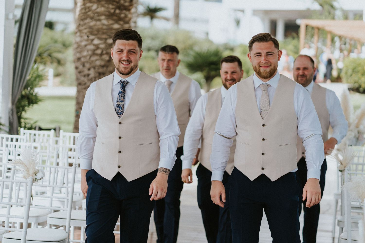 Groomsmen in tan vests and white shirts posing on a tropical venue walkway.