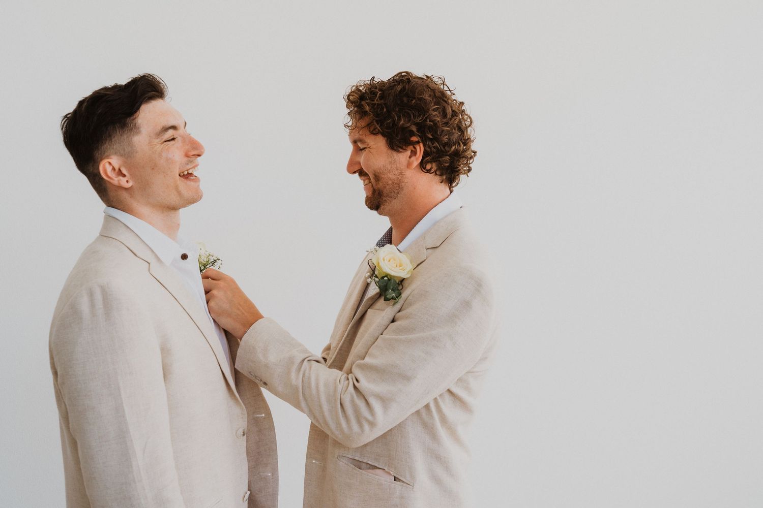 Two groomsmen in beige suits share a joyful moment while adjusting boutonnieres.