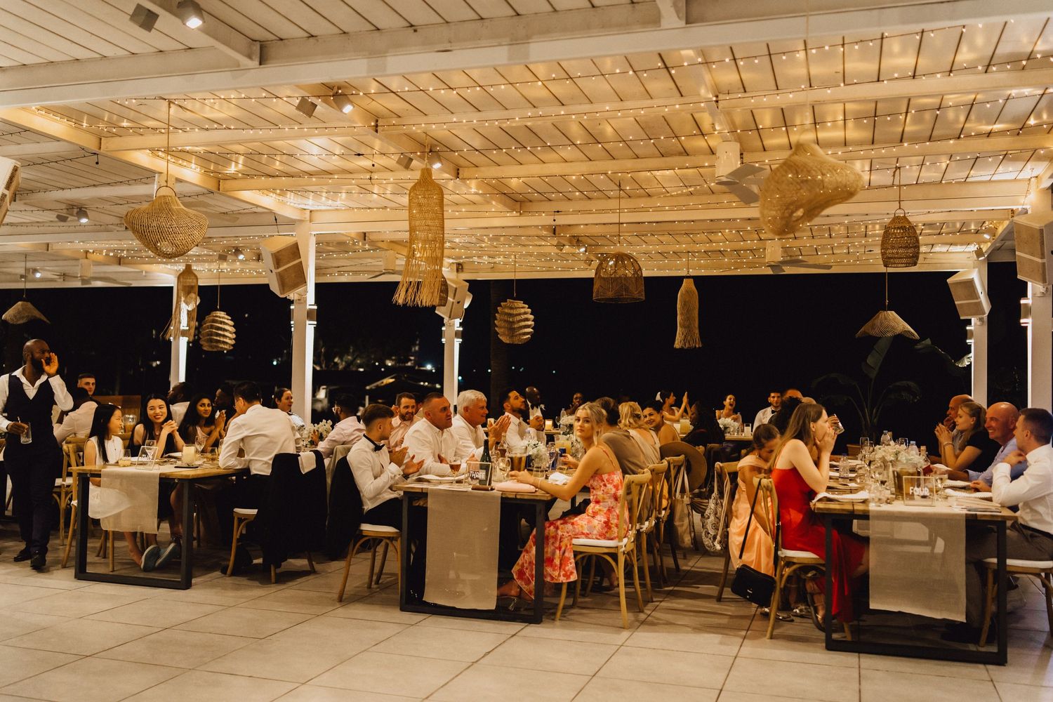 Wedding reception venue decorated with string lights and pendant lamps features tables filled with guests enjoying dinner.