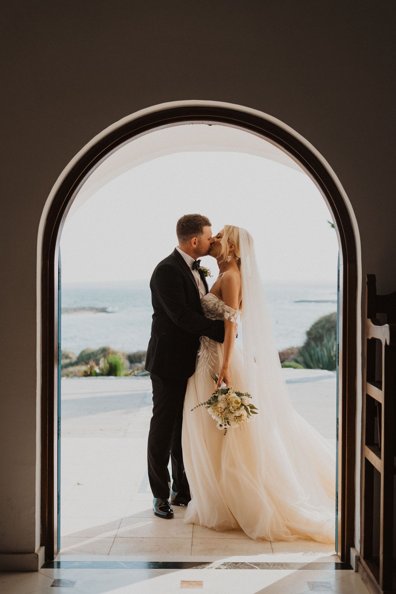 Silhouetted wedding couple kisses in arched doorway overlooking coastal view.