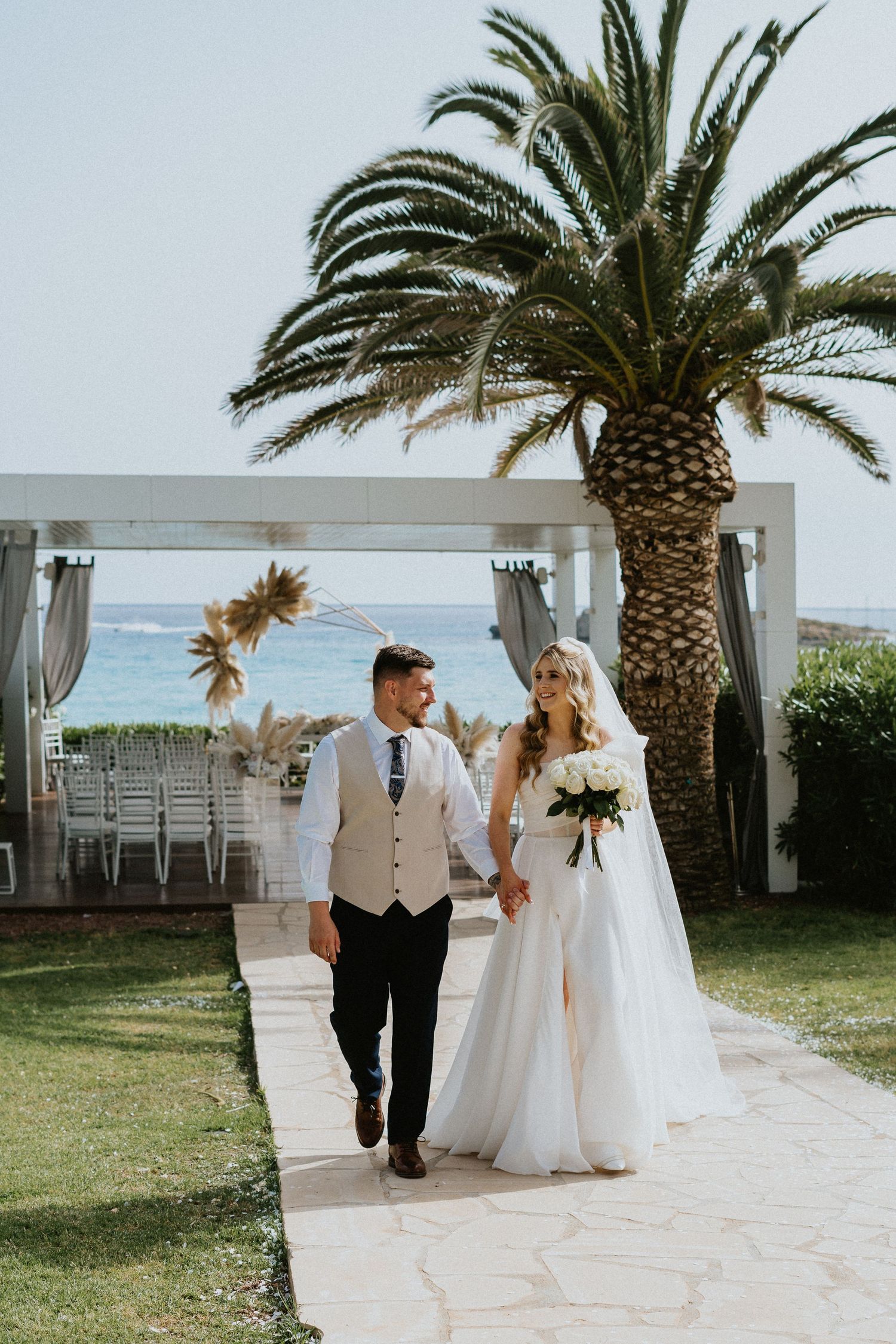 A couple walks hand in hand after their outdoor wedding ceremony under a palm tree by the ocean.