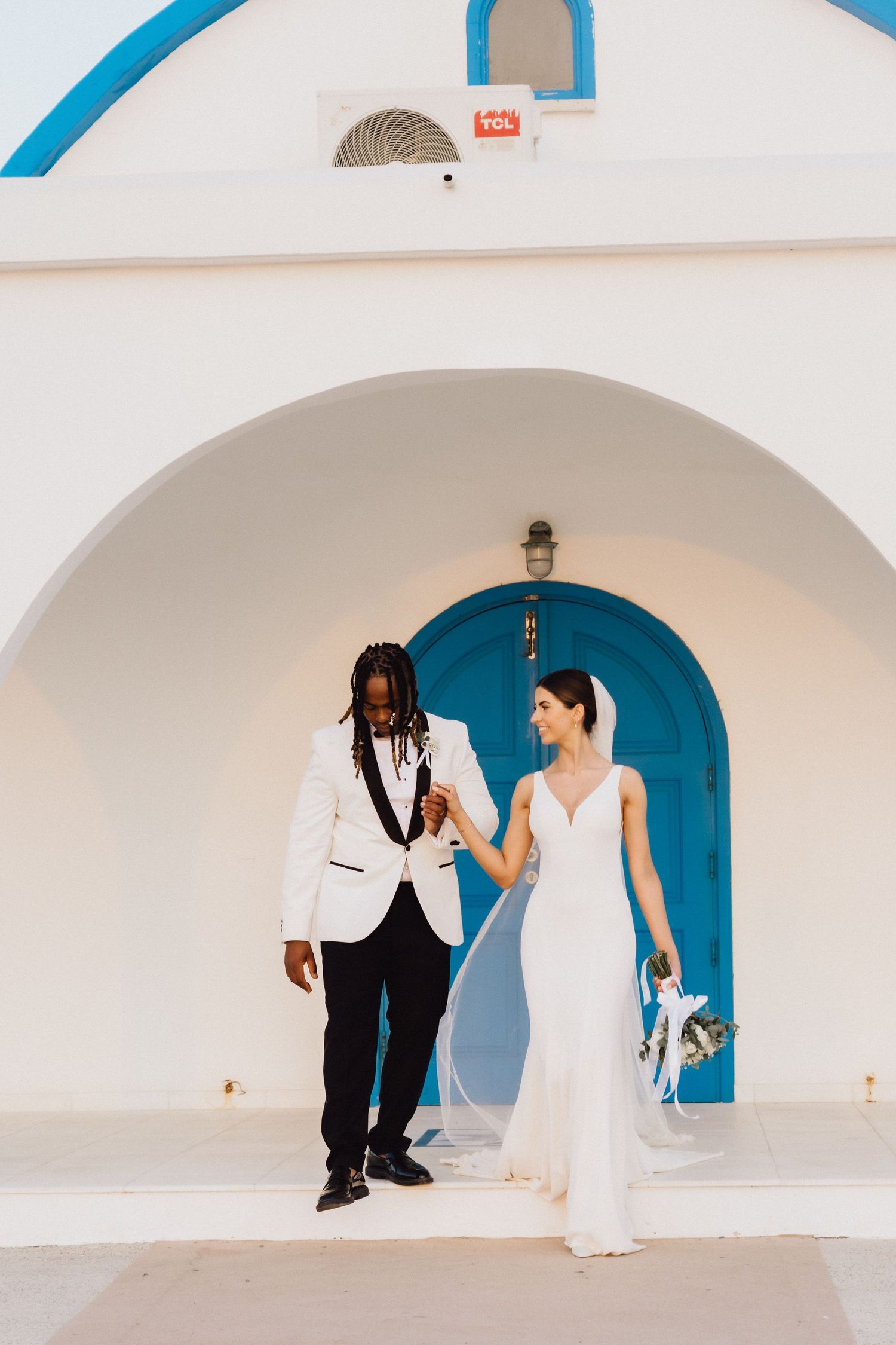 Joyful wedding couple poses against a white Mediterranean building with blue door.