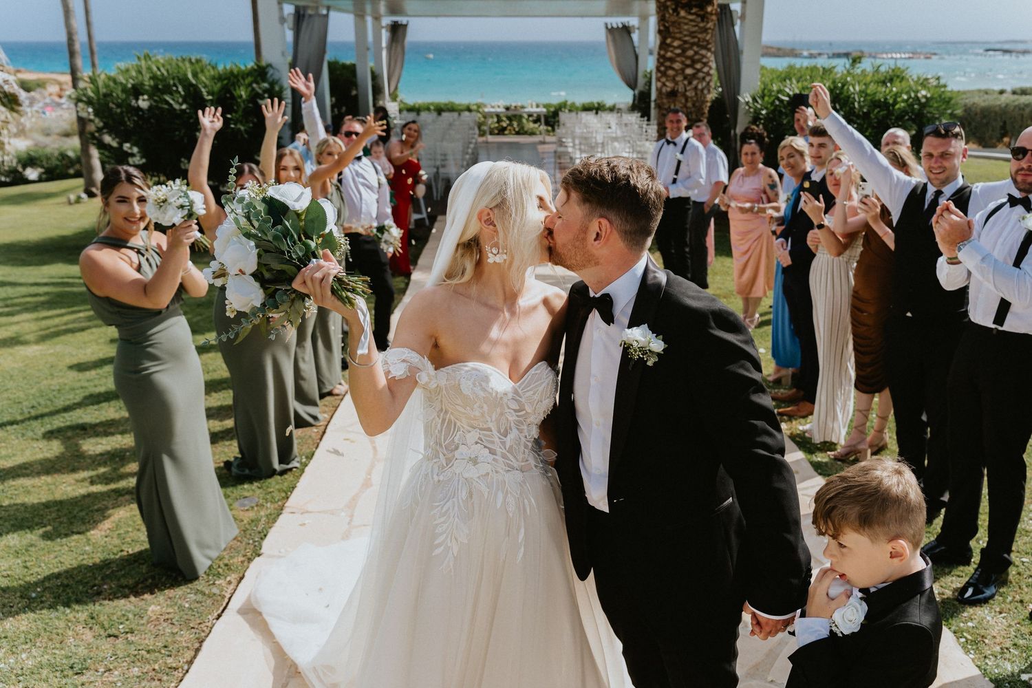 Wedding party cheers as newlyweds share a moment under a palm-lined beachfront gazebo.
