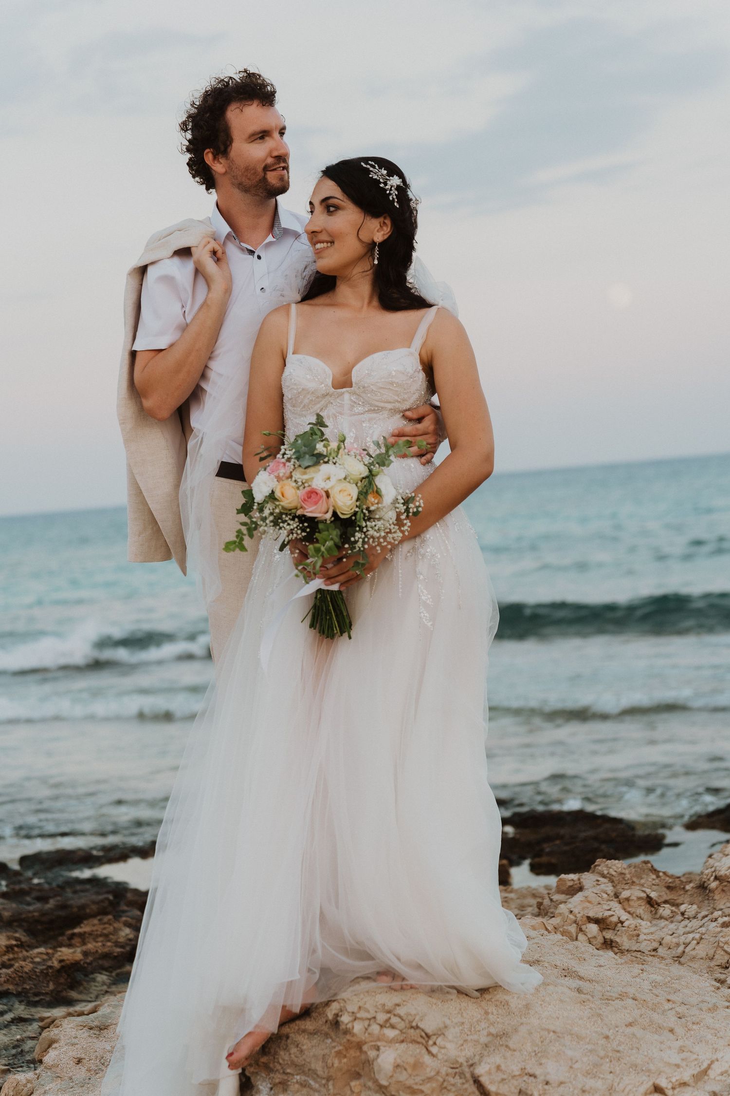 A romantic beach wedding photo of newlyweds standing on rocky shore with ocean backdrop and bridal bouquet.
