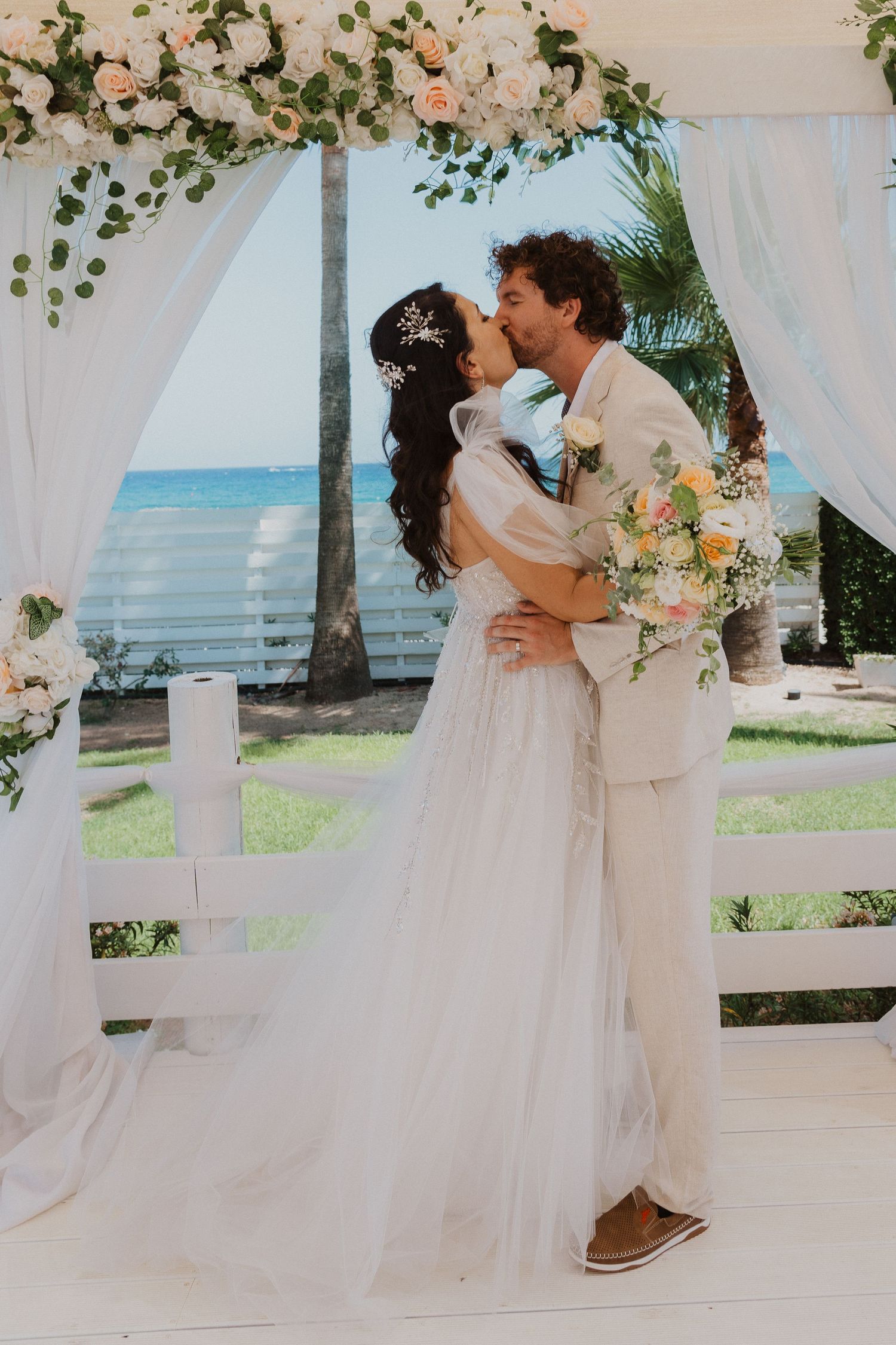 A romantic beach wedding ceremony with a couple kissing under a floral arch with white drapes and ocean views.