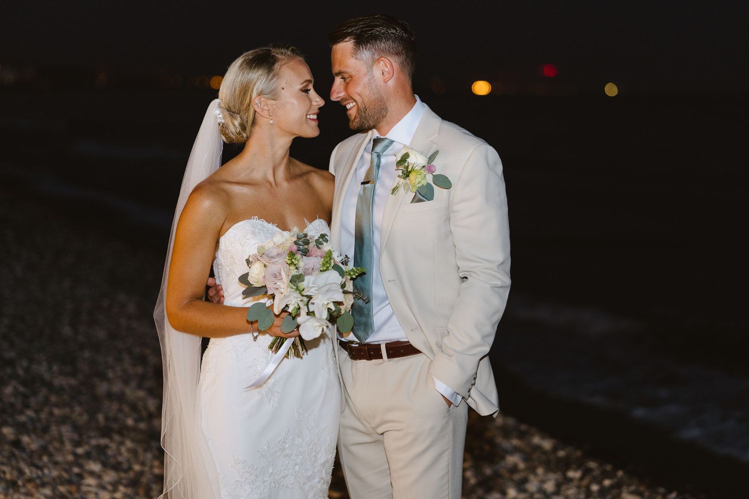 A romantic wedding couple shares a tender moment on a beach at night, with the bride holding white flowers.