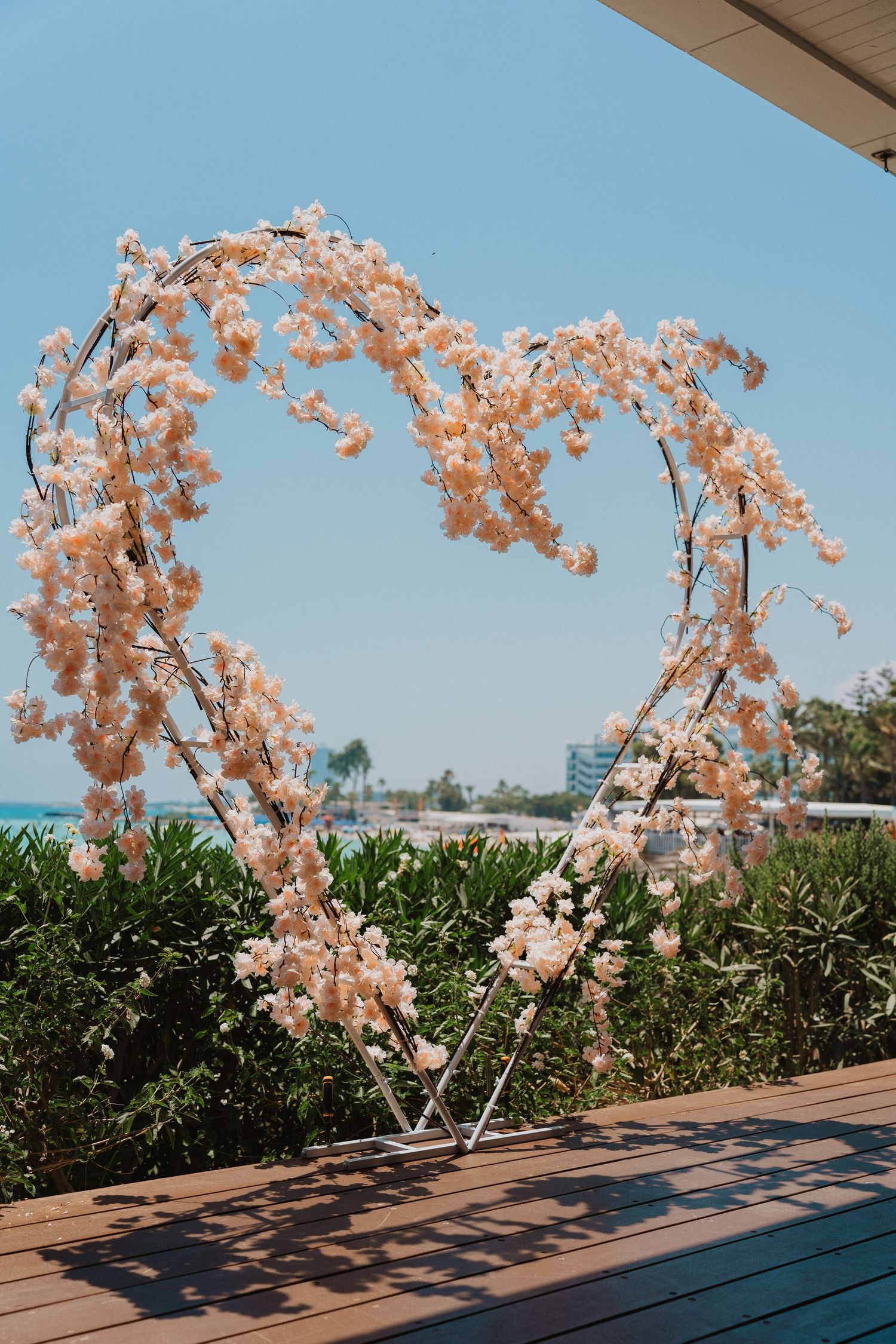 Pink floral heart-shaped wedding arch against blue sky and ocean view.