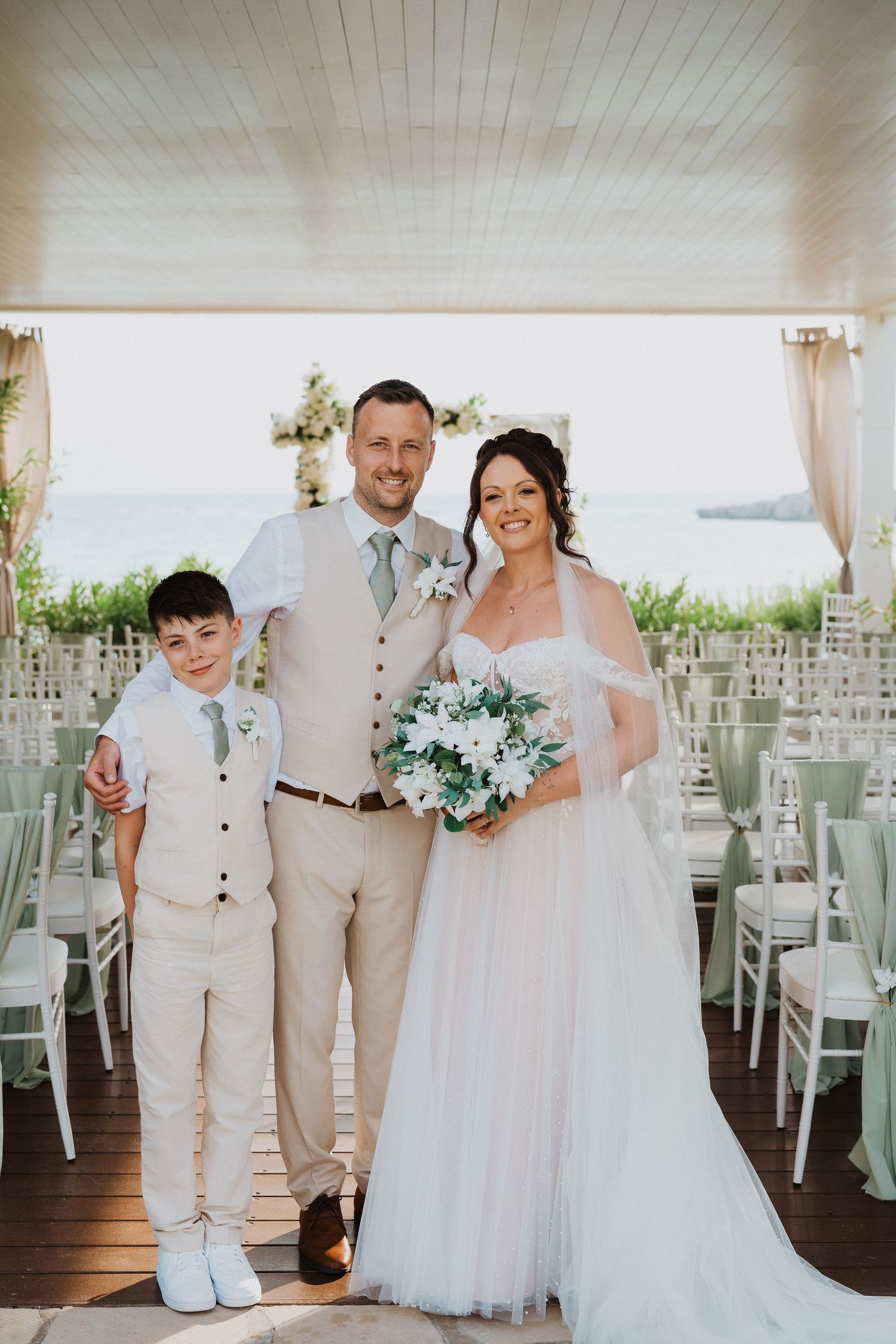 A wedding couple and young boy in light-colored attire pose together at an elegant waterfront venue with white chairs.