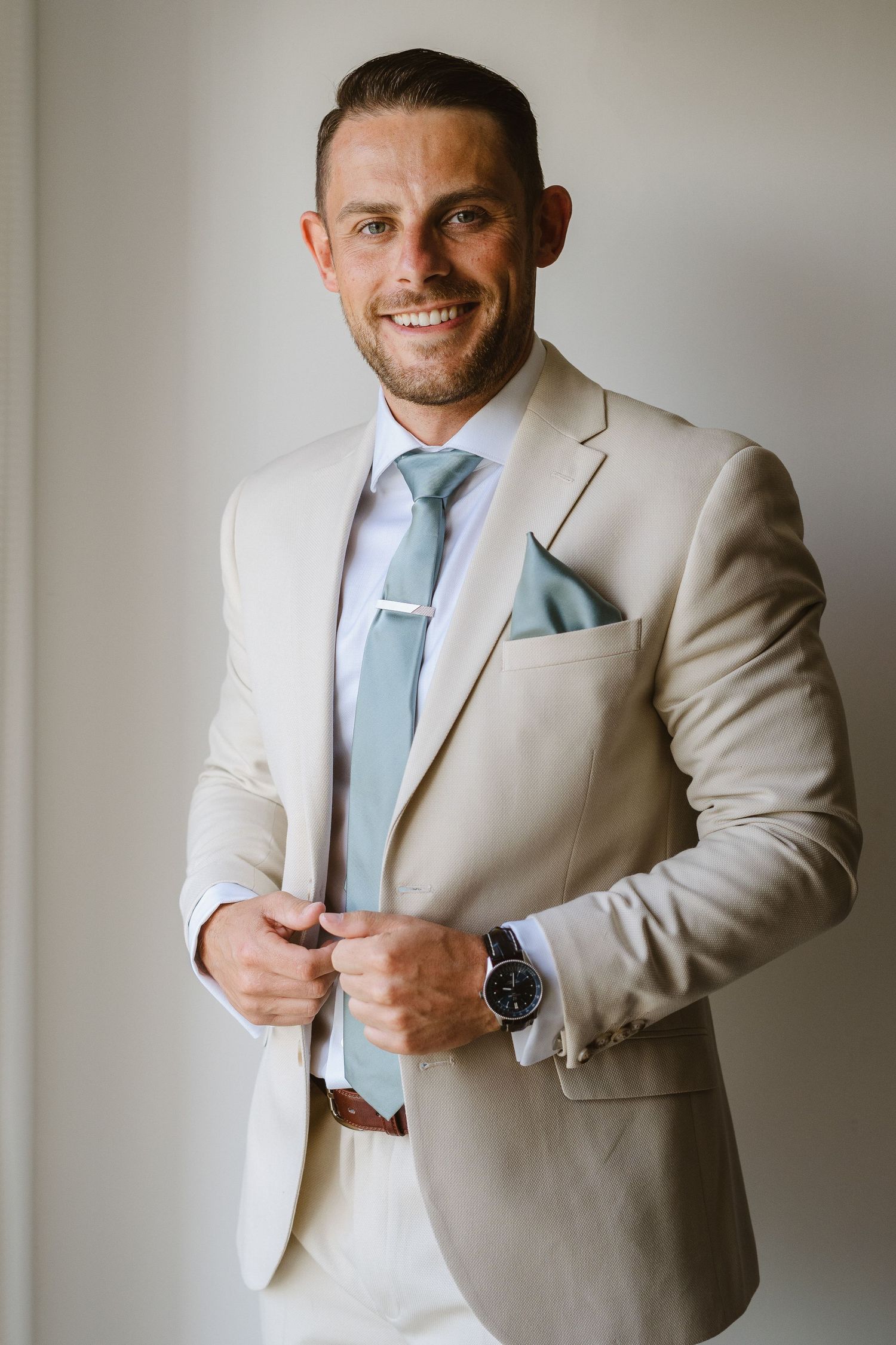 Well-dressed man in cream colored suit and light blue tie adjusts his jacket with a bright smile.